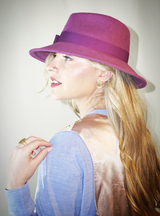 A woman poses in a left side view, wearing the handcrafted Amaryllis fedora hat in luxury purple wool felt with a purple Italian cotton grosgrain ribbon bow. An ideal choice for everyday luxury elegance. Bespoke Couture Millinery by Anna Xaniotaki