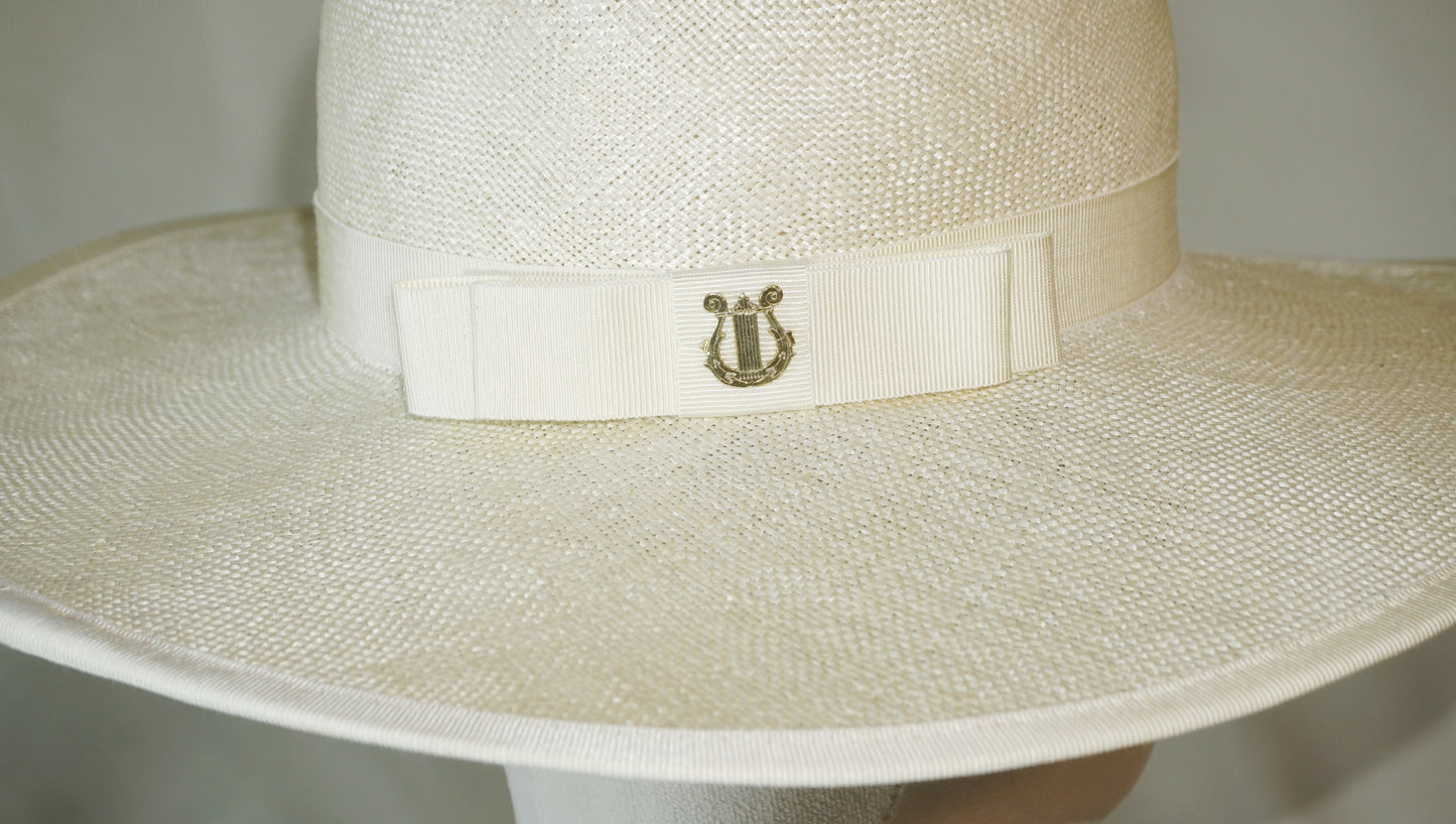Macro detail shot of The Hera luxury handcrafted wide-brim ramie-sisal straw hat, highlighting the 14ct gold-plated lyre jewel carved with Minoan sea lilies, set on a light cream Italian cotton grosgrain band and bow with a hand-basted and machine-stitched grosgrain bound edge. Bespoke Couture Millinery by Anna Xaniotaki