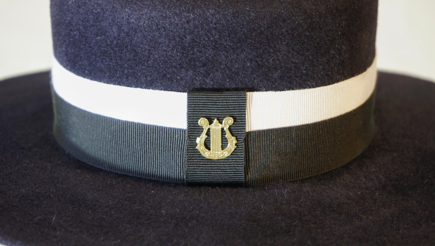 Macro detail shot of the handcrafted Kalypso boater hat, highlighting the signature 14ct gold-plated sterling silver lyre jewel on an ivory and black Italian cotton grosgrain band against the luxury navy blue velour rabbit fur felt texture. Bespoke Couture Millinery by Anna Xaniotaki