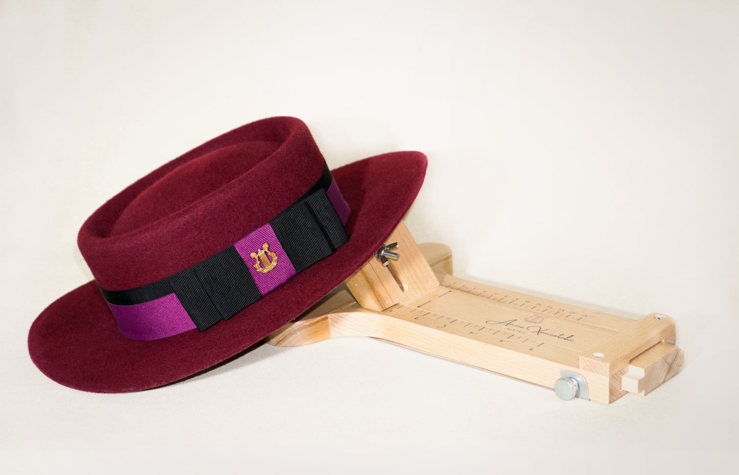 Scarlet boater hat in burgundy fine wool felt with black and orchid purple Italian cotton grosgrain ribbon band, double bow and 14ct gold-plated sterling silver lyre jewel by Anna Xaniotaki ATHENA, displayed upon a round jack millinery toll