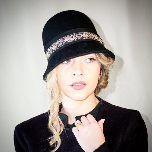 Woman wearing the Vintage Whisper black wool felt cloche hat with a gold lyre emblem, featuring an itallian black grosgrain ribbon and a floral woven ribbon, front view, handcrafted by Anna Xaniotaki.