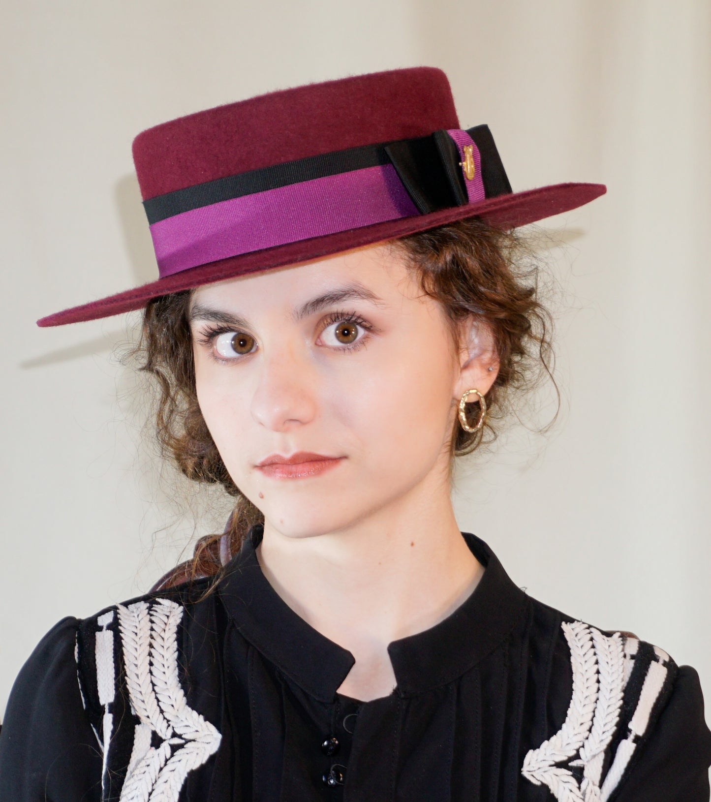 Left angle front view - woman wearing Scarlet Ode burgundy boater hat with double bow and golden lyre detail by Anna Xaniotaki