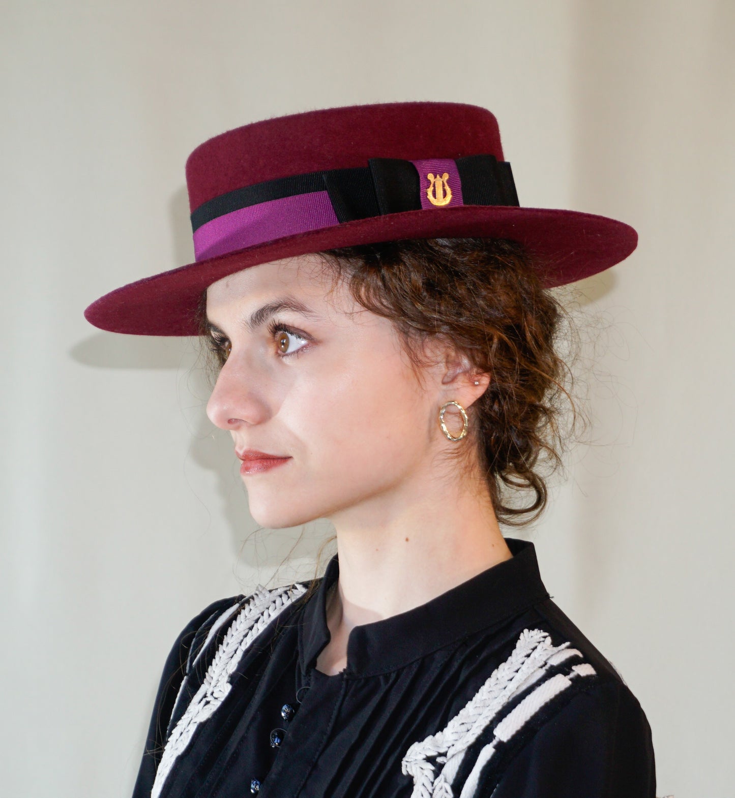Left side view - woman wearing Scarlet Ode burgundy boater hat with golden lyre detail by Anna Xaniotaki