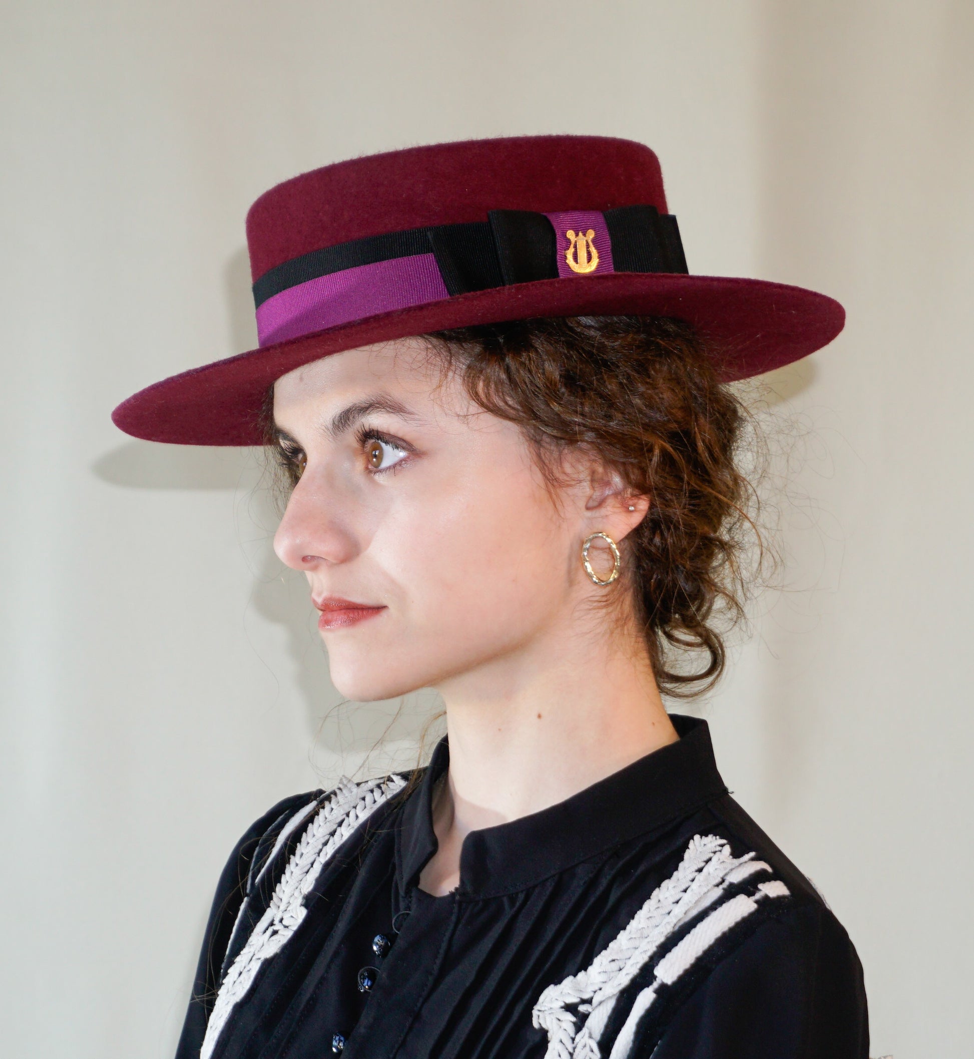 Left side view - woman wearing Scarlet Ode burgundy boater hat with golden lyre detail by Anna Xaniotaki