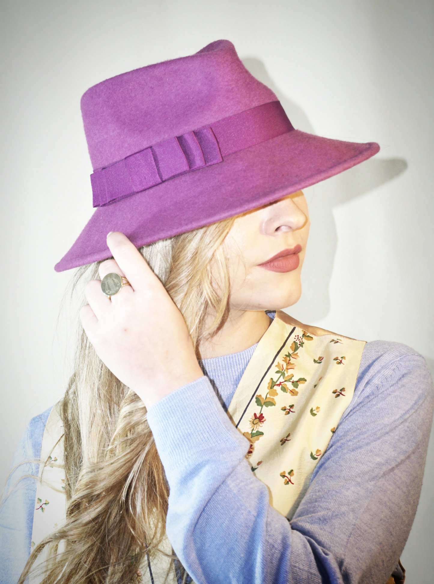 A woman poses in a three-quarter front right view, wearing the handcrafted Amaryllis fedora hat in luxury purple wool felt with a purple Italian cotton grosgrain ribbon bow. An ideal choice for everyday luxury elegance. Bespoke Couture Millinery by Anna Xaniotaki