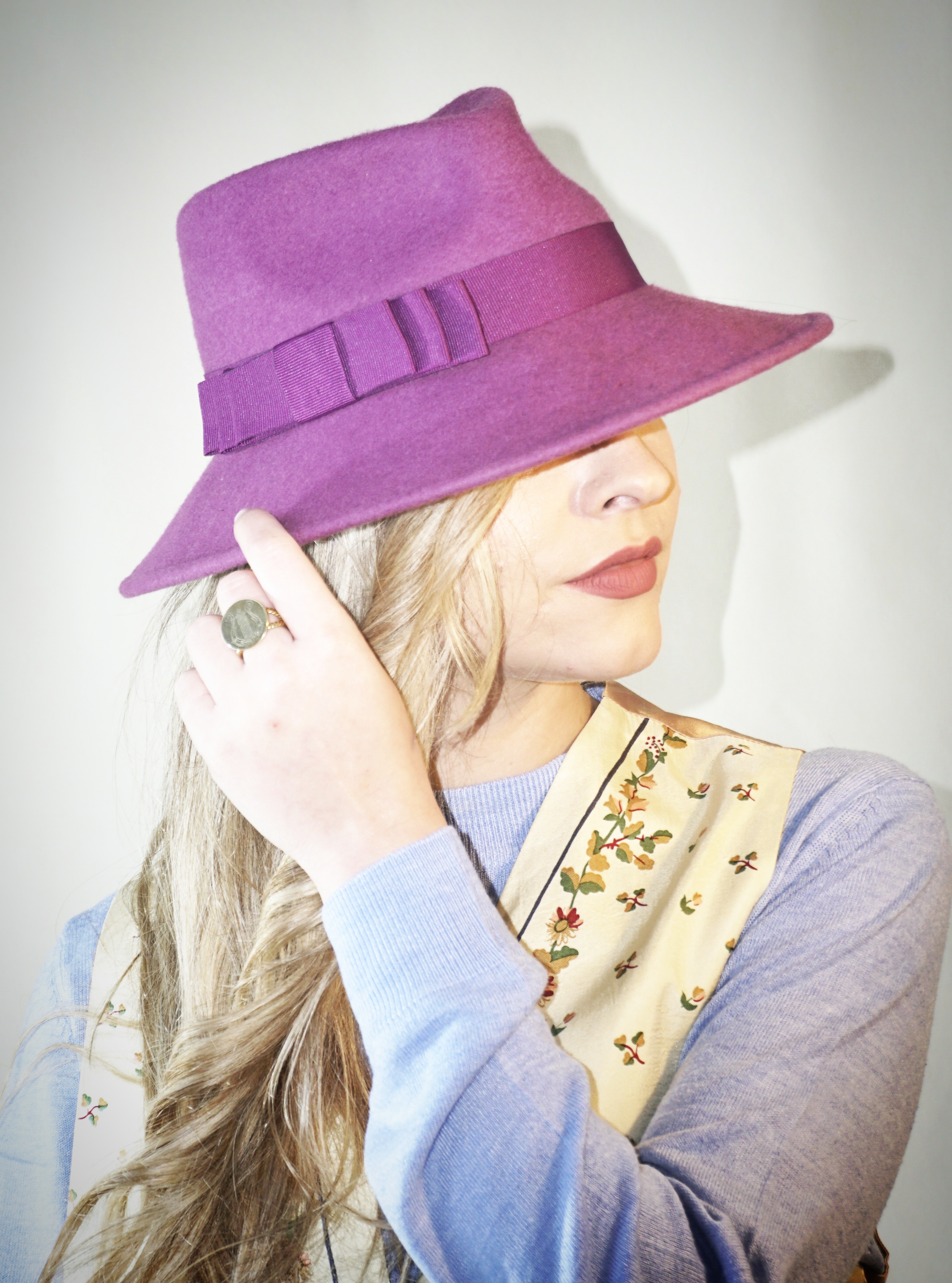 A woman poses in a three-quarter front right view, wearing the handcrafted Amaryllis fedora hat in luxury purple wool felt with a purple Italian cotton grosgrain ribbon bow. An ideal choice for everyday luxury elegance. Bespoke Couture Millinery by Anna Xaniotaki