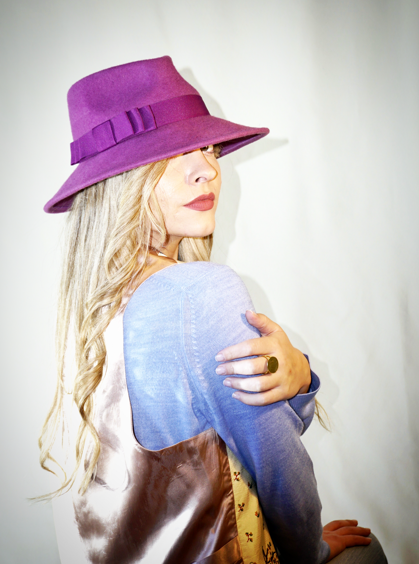 A woman sits with her back turned, glancing forward over her right shoulder, wearing the handcrafted Amaryllis fedora hat in luxury purple wool felt with a purple Italian cotton grosgrain ribbon bow. An ideal choice for everyday luxury elegance. Bespoke Couture Millinery by Anna Xaniotaki