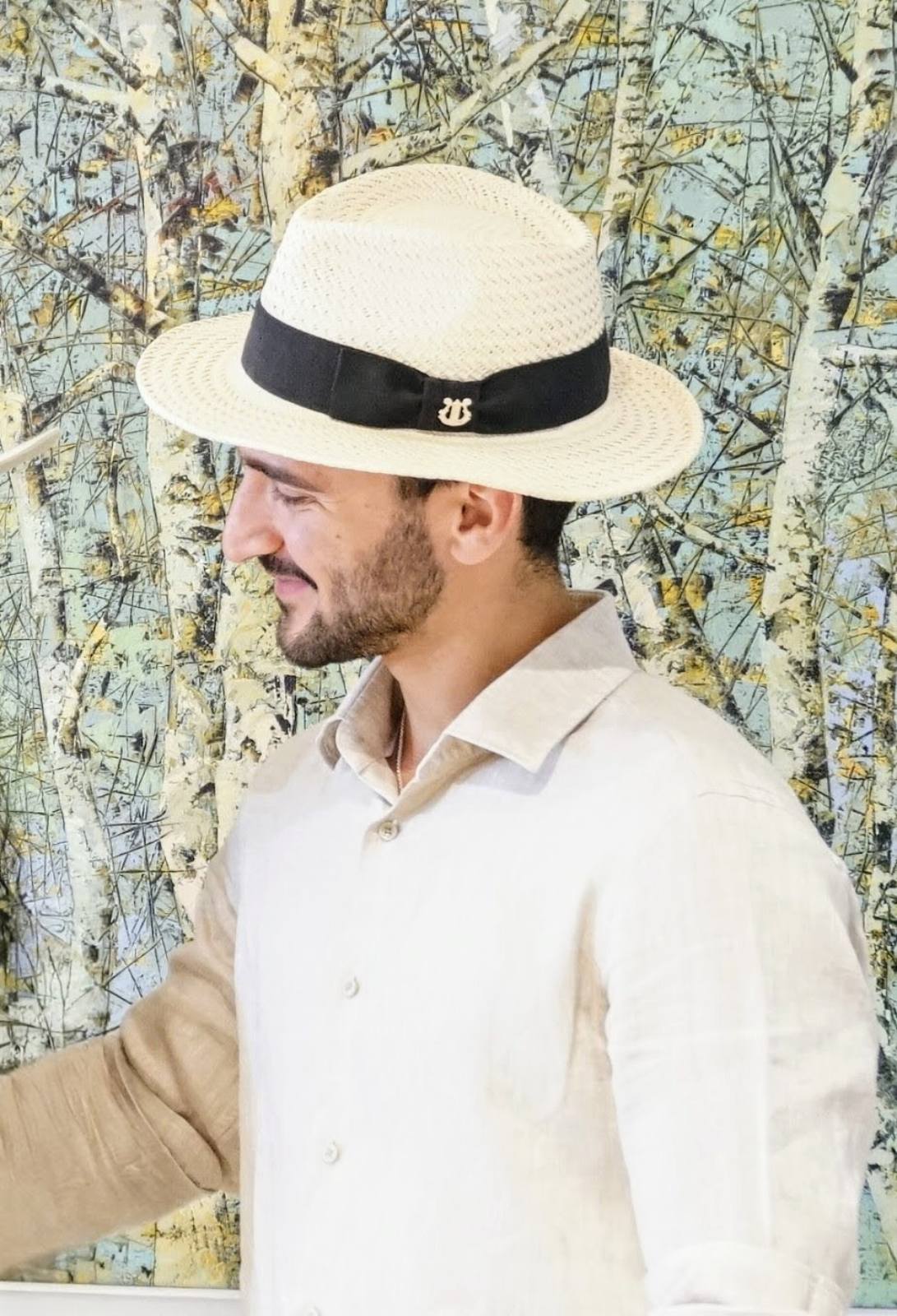 Man wearing the André fedora straw hat in ivory with black Italian cotton grosgrain band and bow and a 925 sterling silver lyre jewel,             
Left side view. Custom made-to-order fedora for summer special occasions, garden parties, vacations or for everyday man's elegance. Bespoke couture millinery by Anna Xaniotaki