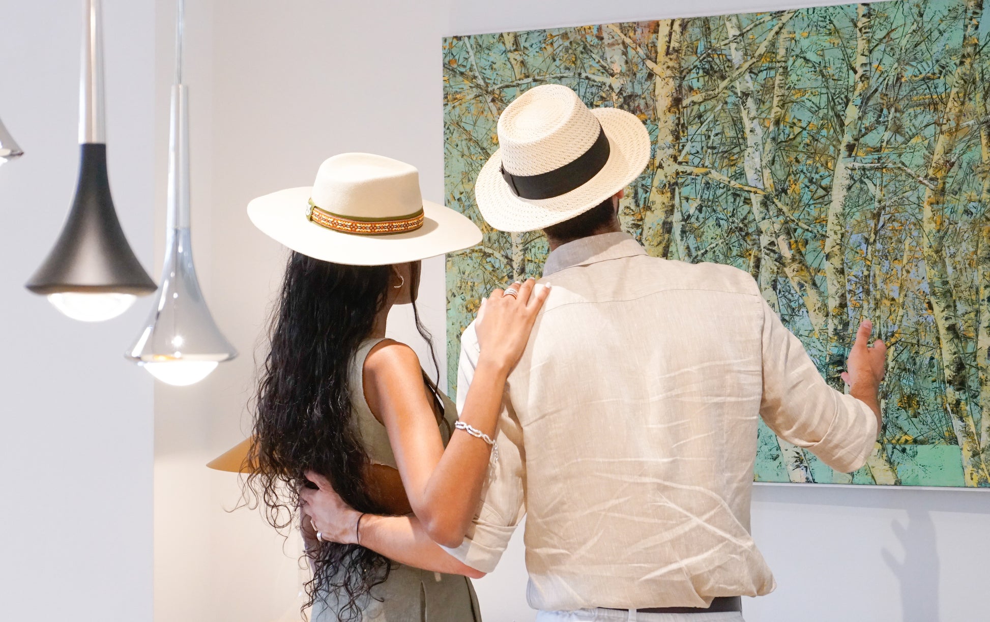 Man wearing the André fedora straw hat in ivory with black Italian cotton grosgrain band, bow and 14ct gold-plated sterling silver lyre jewel, looking at a woman in a light cream fedora, back side view. Custom made-to-order fedora for summer special occasions, garden parties, vacations or for everyday man's elegance. Bespoke couture millinery by Anna Xaniotaki