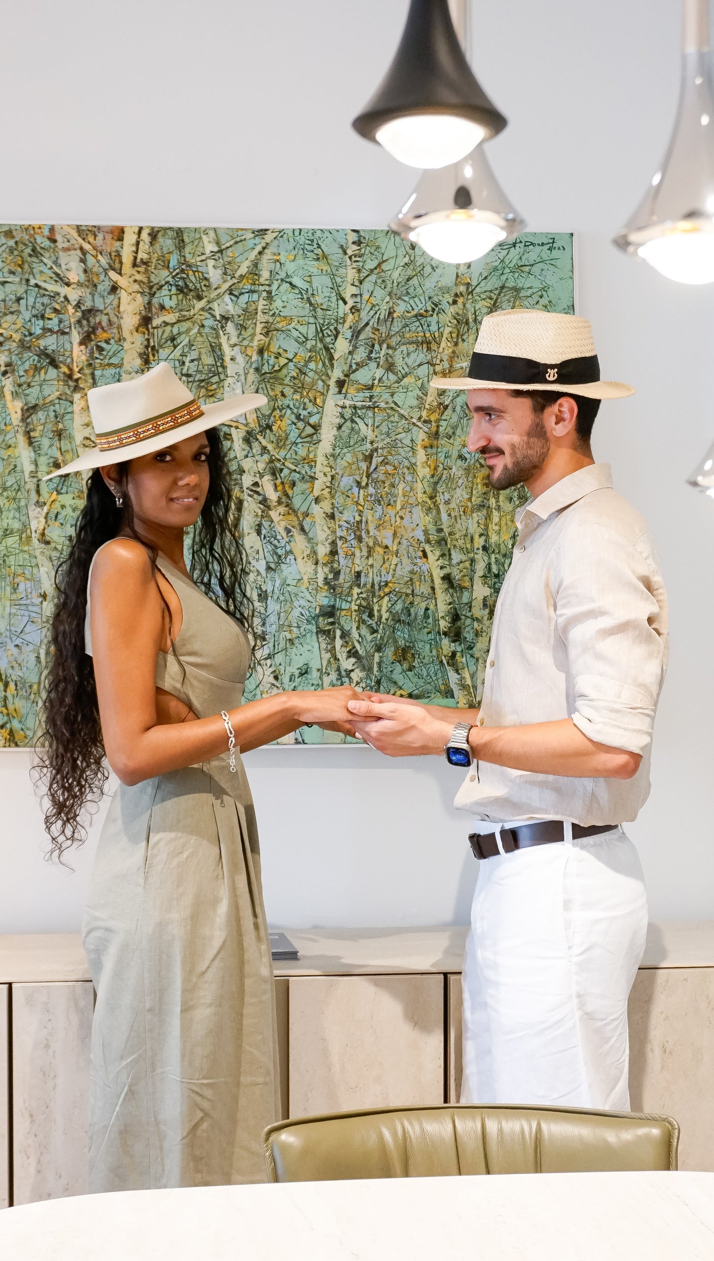 Man wearing the André fedora straw hat in ivory with black Italian cotton grosgrain band, bow and 14ct gold-plated sterling silver lyre jewel, looking at a woman in a light cream fedora and touching her hands, right side view. Custom made-to-order fedora for summer special occasions, garden parties, vacations or for everyday man's elegance. Bespoke couture millinery by Anna Xaniotaki