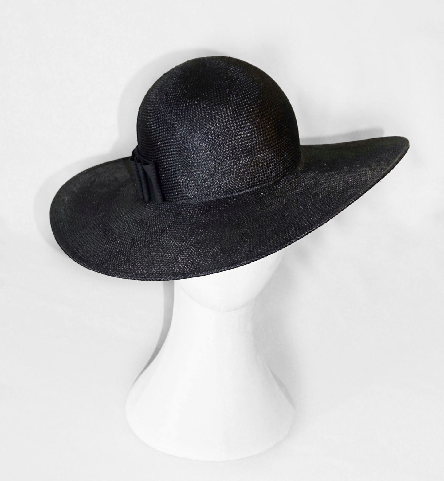 The handcrafted Auréole Élégante wide-brim visca straw hat in black, featuring a glimpse of the tailored black Italian cotton grosgrain bow extending to the back and the signature 14ct gold-plated sterling silver lyre jewel hidden on the right back side, displayed on a white mannequin stand — front view. Bespoke Couture Millinery by Anna Xaniotaki.Xaniotaki 