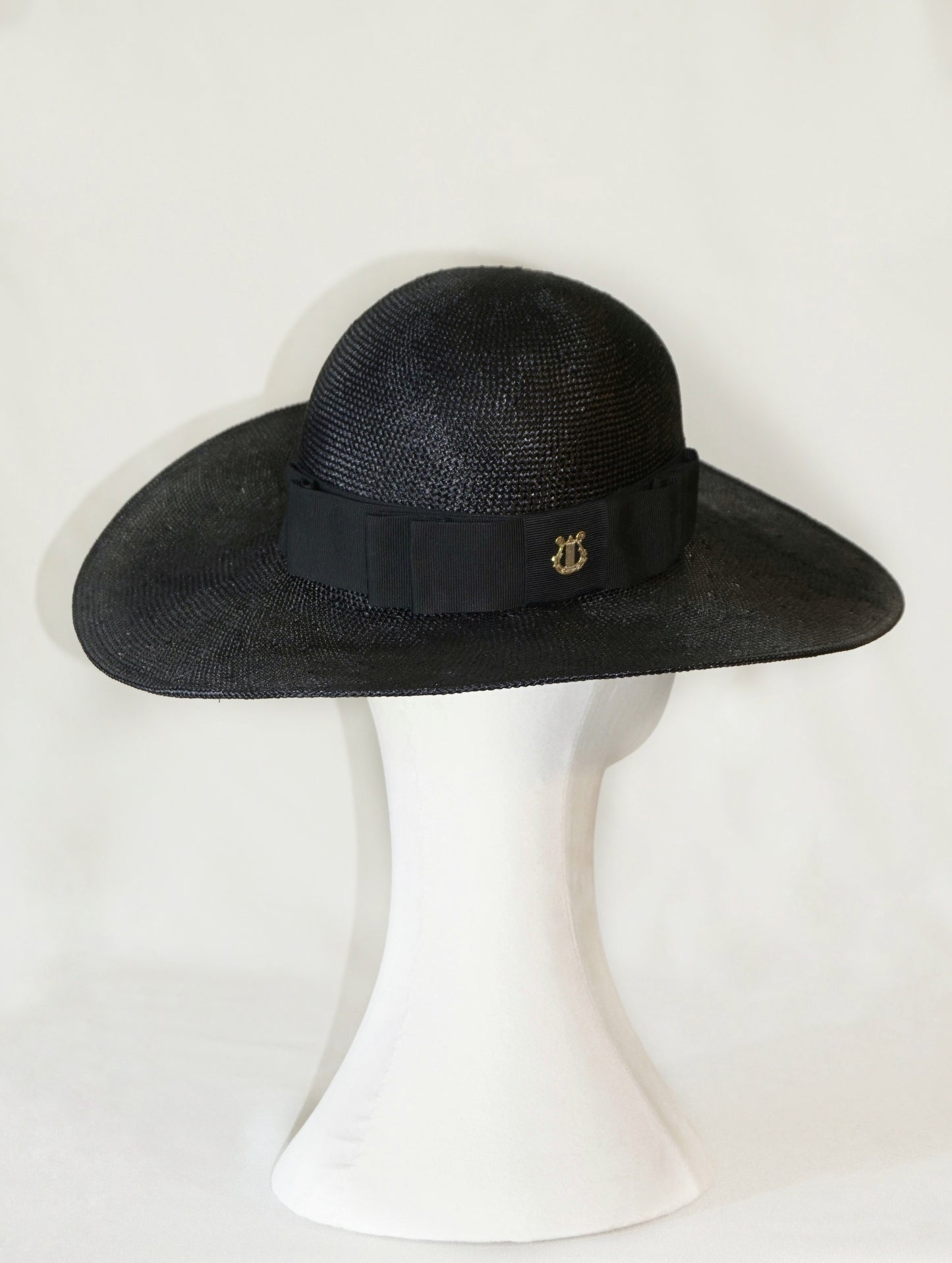 The luxury Auréole Élégante wide-brim visca straw hat in black, featuring a tailored black Italian cotton grosgrain bow and the signature 14ct gold-plated sterling silver lyre jewel, displayed on a white mannequin stand — three-quarter back right view. Bespoke Couture Millinery by Anna Xaniotaki.