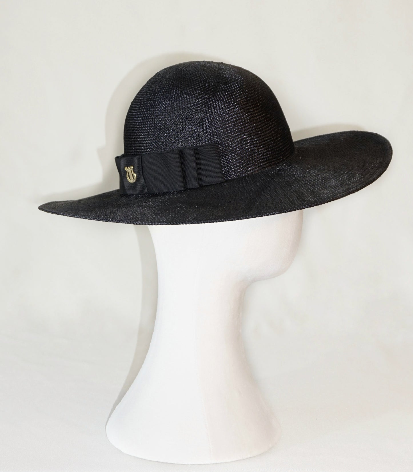 The handcrafted Auréole Élégante wide-brim visca straw hat in black, featuring a tailored black Italian cotton grosgrain bow and the signature 14ct gold-plated sterling silver lyre jewel, displayed on a white mannequin stand — right side view. Bespoke Couture Millinery by Anna Xaniotaki.
