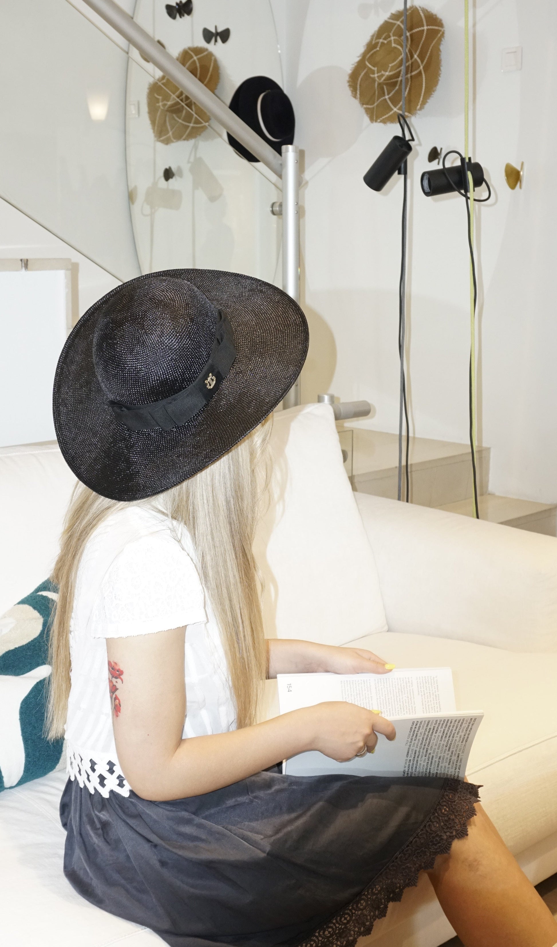 A woman sits on a couch reading a book in a three-quarter back view, wearing the handcrafted Auréole Élégante wide-brim visca straw hat in black with a tailored black Italian cotton grosgrain bow and the signature 14ct gold-plated sterling silver lyre jewel, against an ivory couch and atelier interior background. A statement hat for Royal Ascot, race days, couture weddings, gallery openings, garden & cocktail parties, and everyday luxury elegance. Bespoke Couture Millinery by Anna Xaniotaki.
