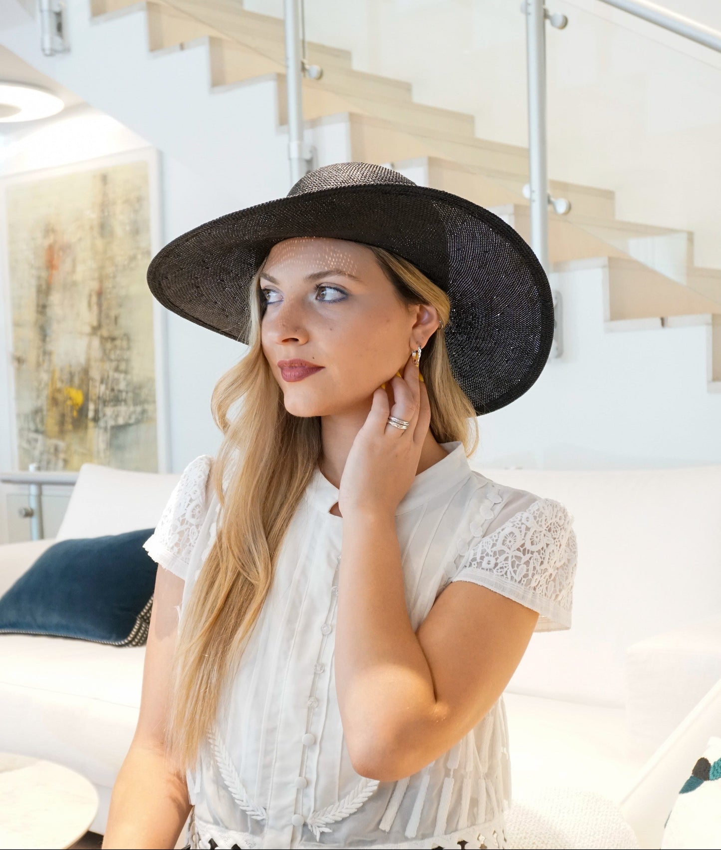 A woman looks away in a three-quarter front left view, wearing the handcrafted Auréole Élégante wide-brim visca straw hat in black with a tailored black Italian cotton grosgrain bow and the signature 14ct gold-plated sterling silver lyre jewel hidden on the right back side, against an ivory couch and atelier interior with a modern staircase in the background. A statement hat for Royal Ascot, race days, couture weddings, and everyday luxury elegance. Bespoke Couture Millinery by Anna Xaniotaki.
