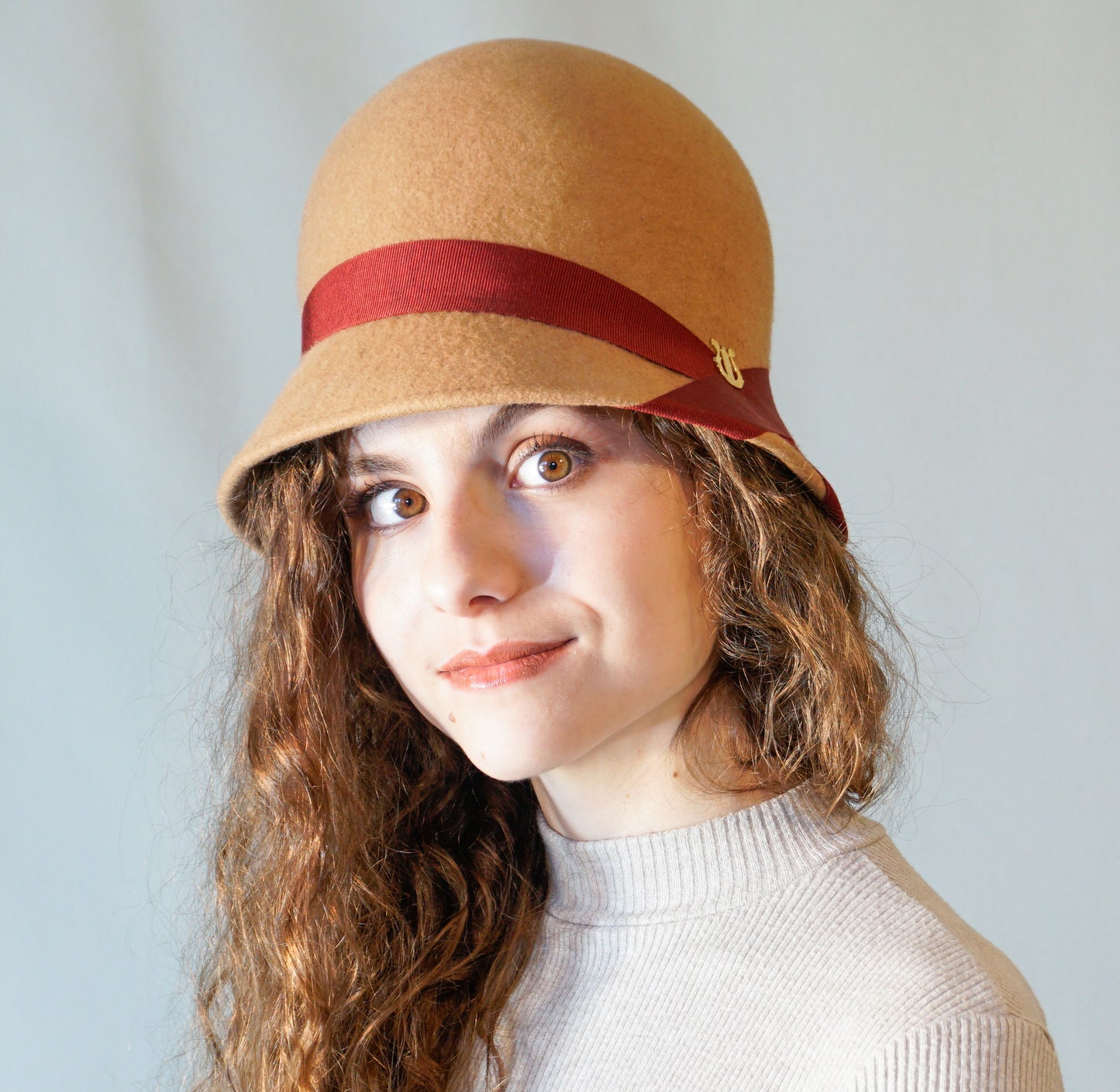 Daize cloche hat in camel wool felt with red ribbon and gold lyre by Anna Xaniotaki - three quarter view