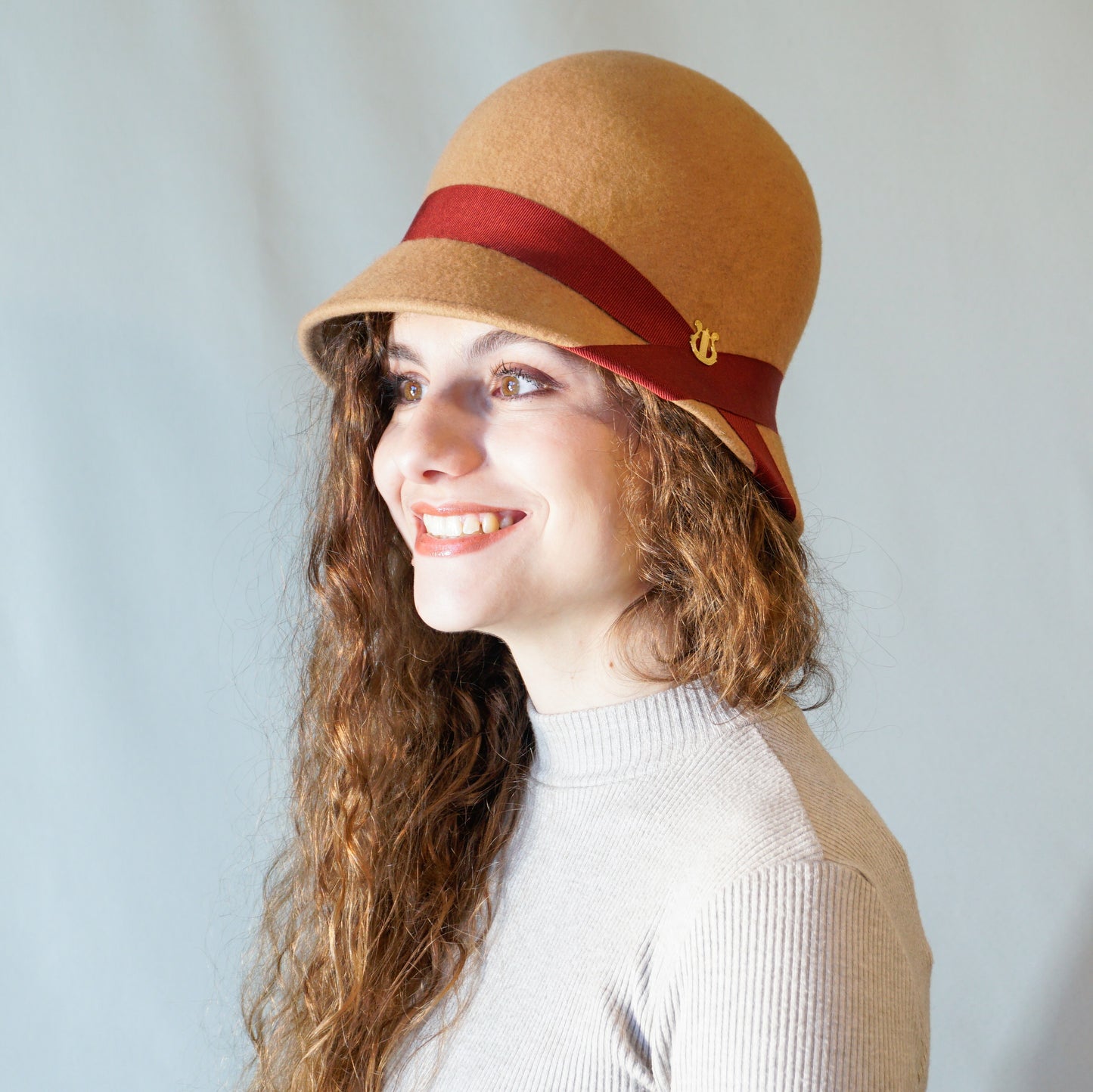 Daize cloche hat in camel wool felt with red ribbon and gold lyre by Anna Xaniotaki - three quarter view