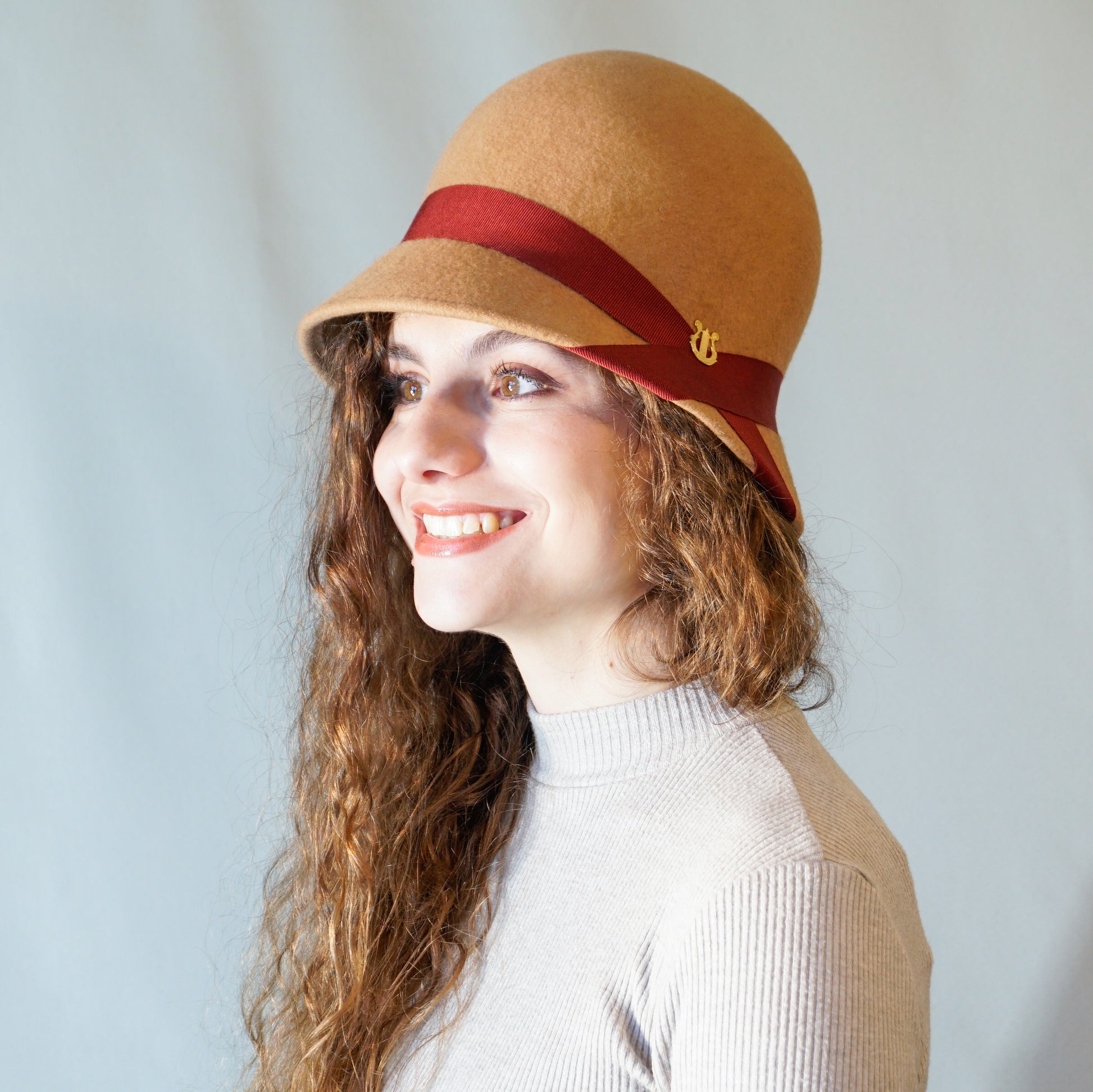 Daize cloche hat in camel wool felt with red ribbon and gold lyre by Anna Xaniotaki - three quarter view