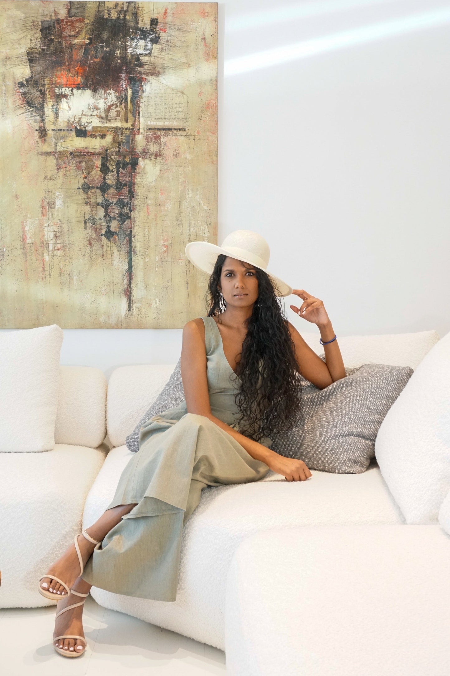 The Hera luxury handcrafted wide-brim ramie-sisal straw hat in champagne, featuring a light cream Italian cotton grosgrain band & a tailored grosgrain bound edge. Shown in a full-length front view on a woman sitting on a couch and touching the brim. An abstract painting and luxury interior in the background. An exquisite Royal Ascot, race day & couture wedding hat or for everyday luxury. Bespoke Couture Millinery by Anna Xaniotaki 