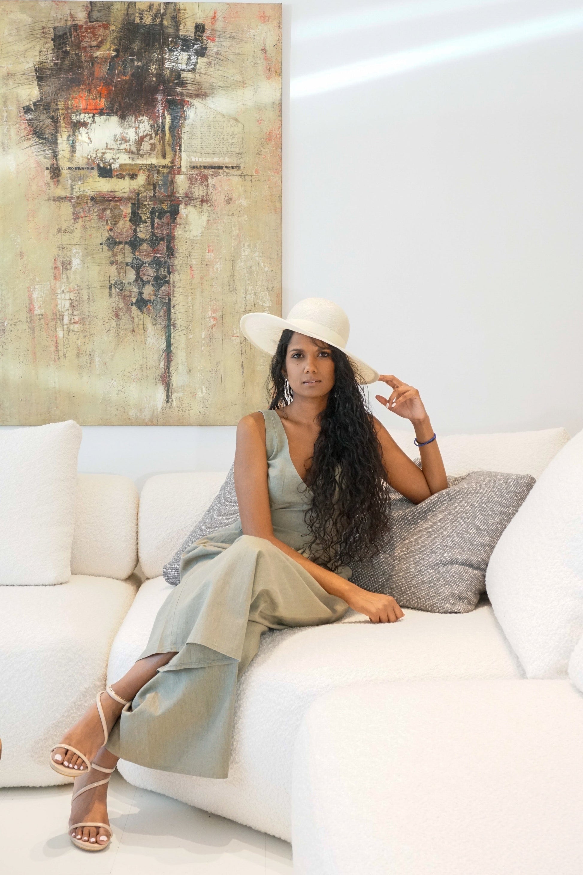 The Hera luxury handcrafted wide-brim ramie-sisal straw hat in champagne, featuring a light cream Italian cotton grosgrain band & a tailored grosgrain bound edge. Shown in a full-length front view on a woman sitting on a couch and touching the brim. An abstract painting and luxury interior in the background. An exquisite Royal Ascot, race day & couture wedding hat or for everyday luxury. Bespoke Couture Millinery by Anna Xaniotaki 