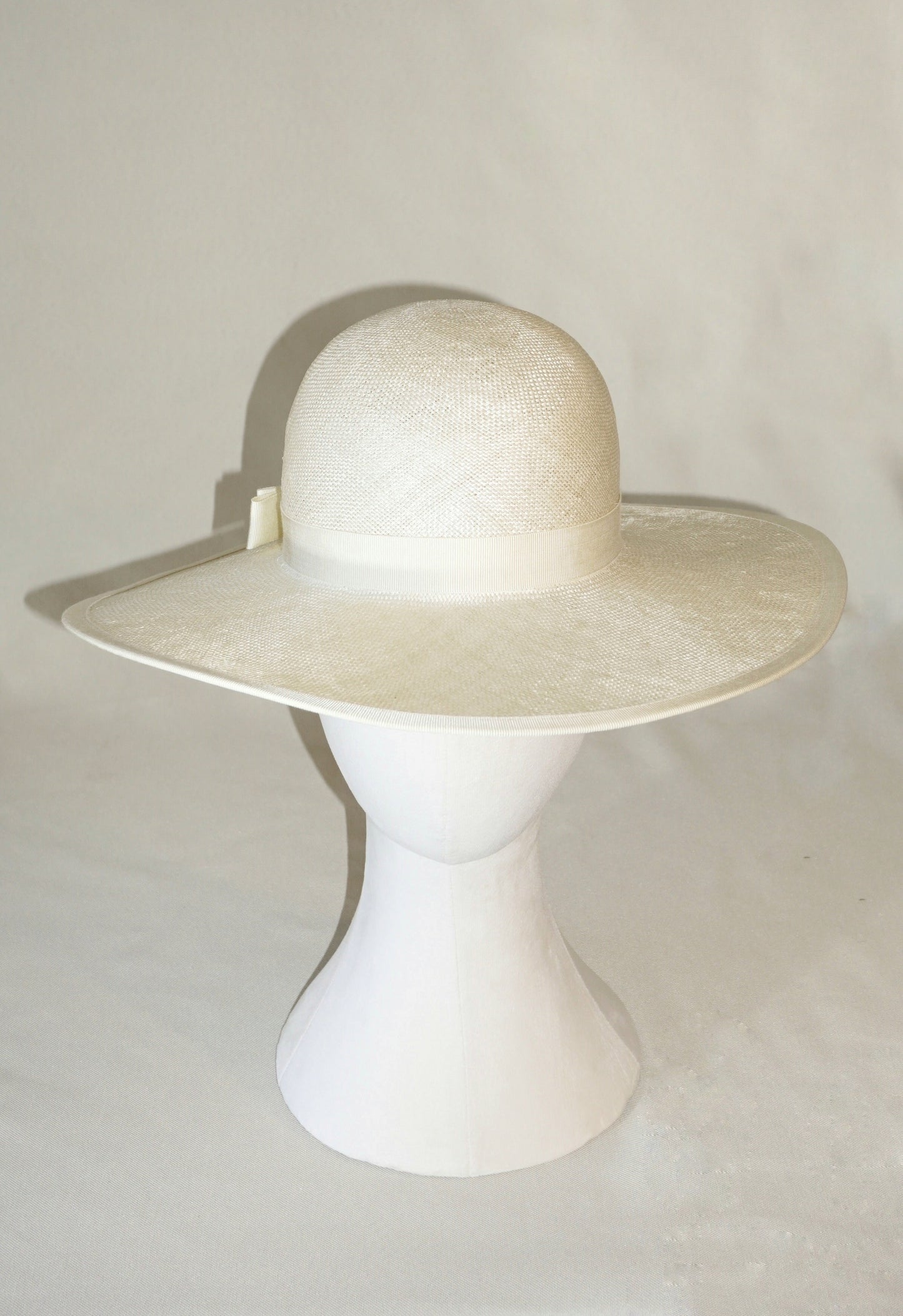  The Hera luxury handcrafted wide-brim ramie-sisal straw hat in champagne, featuring a light cream Italian cotton grosgrain band & a tailored grosgrain bound edge, with the matching bow & 14ct gold-plated lyre jewellery hidden on the three-quarter back right side of the hat. Front view product shot displayed on a white mannequin stand. Bespoke Couture Millinery by Anna Xaniotaki