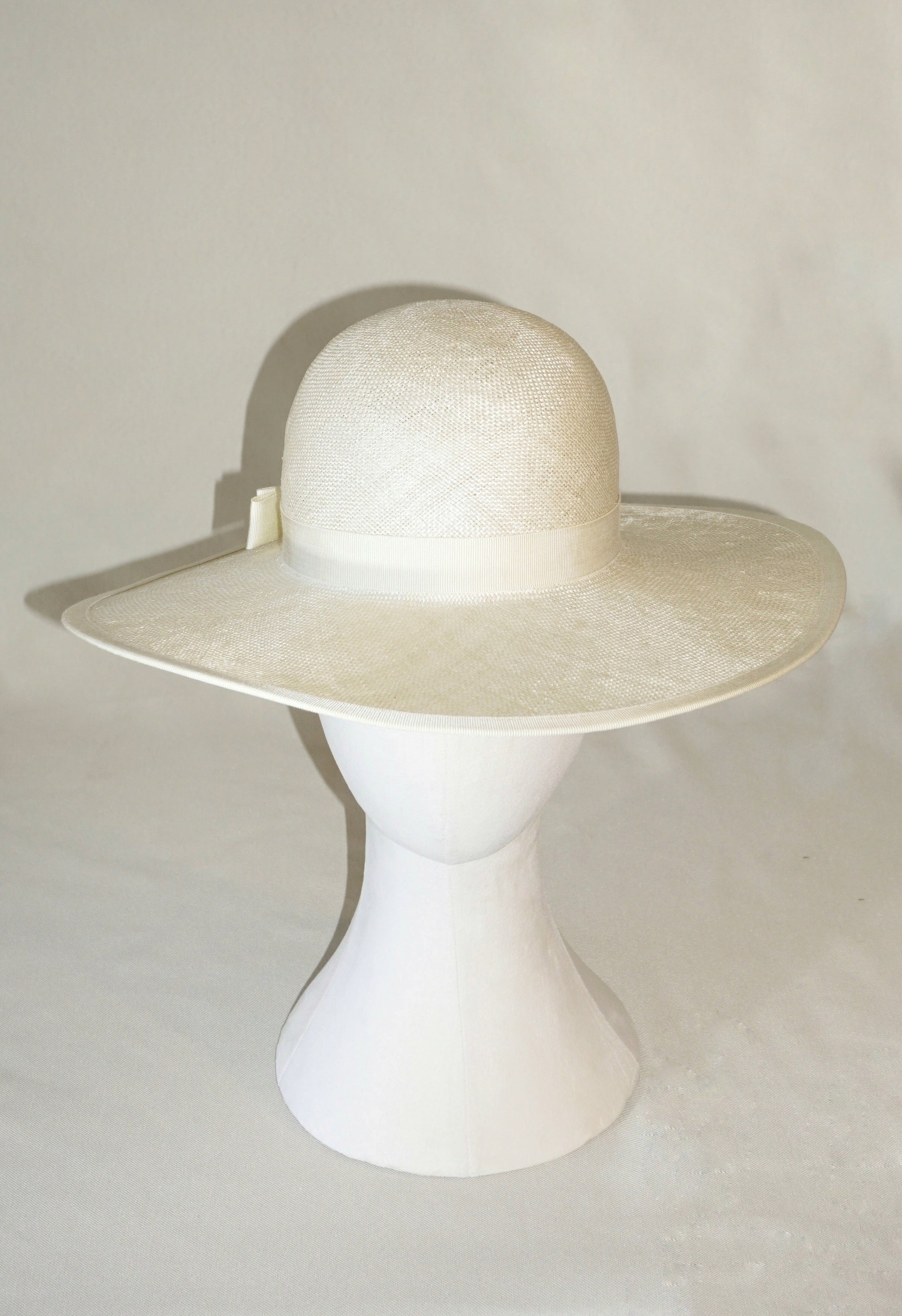  The Hera luxury handcrafted wide-brim ramie-sisal straw hat in champagne, featuring a light cream Italian cotton grosgrain band & a tailored grosgrain bound edge, with the matching bow & 14ct gold-plated lyre jewellery hidden on the three-quarter back right side of the hat. Front view product shot displayed on a white mannequin stand. Bespoke Couture Millinery by Anna Xaniotaki