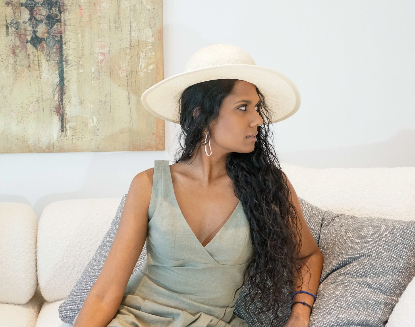 The Hera luxury handcrafted wide-brim ramie-sisal straw hat in champagne, featuring a light cream Italian cotton grosgrain band & a tailored grosgrain bound edge. Shown in a close-up three-quarter front right view on a woman sitting on a couch and looking away. An abstract painting and luxury interior in the background. An exquisite Royal Ascot, race day & couture wedding hat or for everyday luxury. Bespoke Couture Millinery by Anna Xaniotaki 