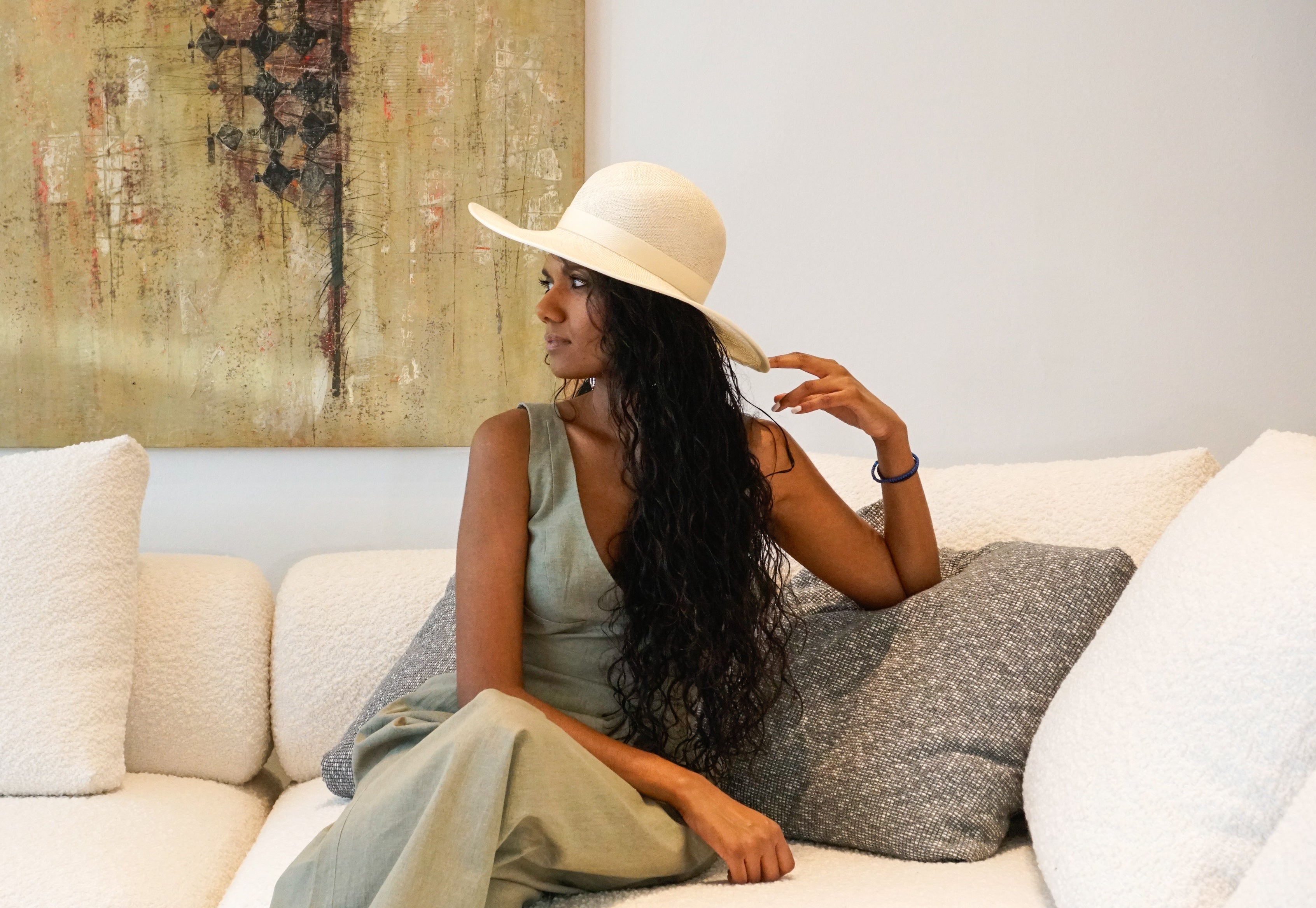 The Hera luxury handcrafted wide-brim ramie-sisal straw hat in champagne, featuring a light cream Italian cotton grosgrain band & a tailored grosgrain bound edge, with the matching bow & signature 14ct gold-plated lyre jewel hidden on the right back side. Shown in a three-quarter length left profile view on a woman sitting on a couch and touching the brim. An exquisite Royal Ascot, race day & couture wedding hat or for everyday luxury. Bespoke Couture Millinery by Anna Xaniotaki