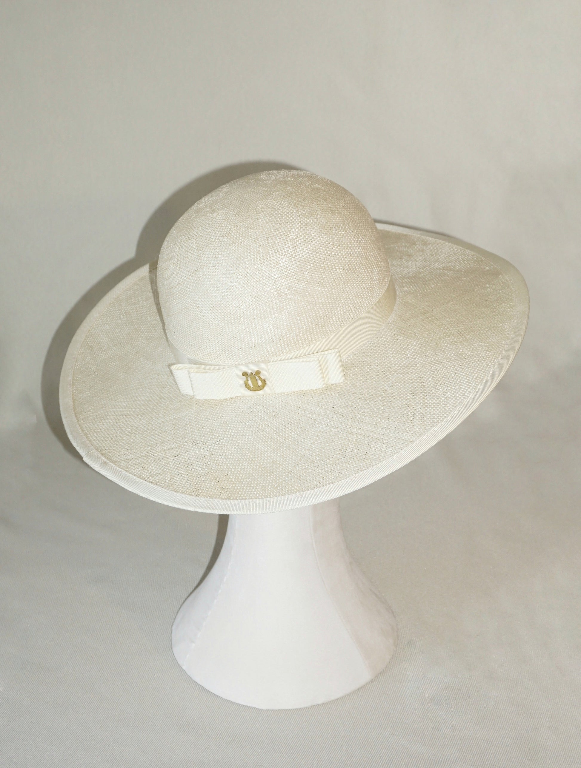 The Hera luxury handcrafted wide-brim ramie-sisal straw hat in champagne, featuring a light cream Italian cotton grosgrain band, a matching bow, a tailored grosgrain bound edge & 14ct gold-plated lyre jewellery. High-angle three-quarter back right view product shot displayed on a white mannequin stand. Bespoke Couture Millinery by Anna Xaniotaki