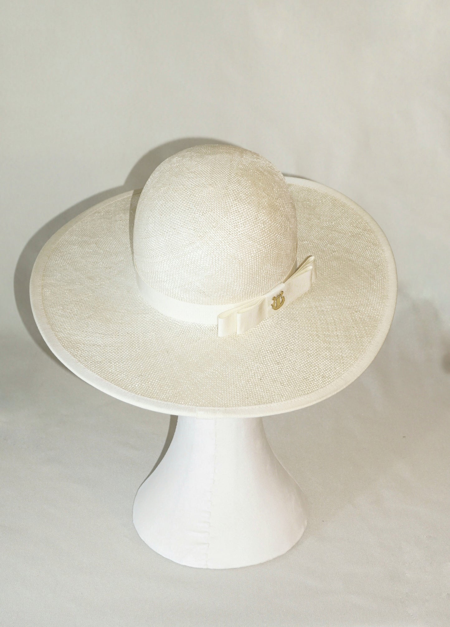 The Hera luxury handcrafted wide-brim ramie-sisal straw hat in champagne, featuring a light cream Italian cotton grosgrain band & a tailored grosgrain bound edge, with the matching bow & 14ct gold-plated lyre jewellery positioned on the three-quarter back right side of the hat. High-angle back view product shot displayed on a white mannequin stand. Bespoke Couture Millinery by Anna Xaniotaki