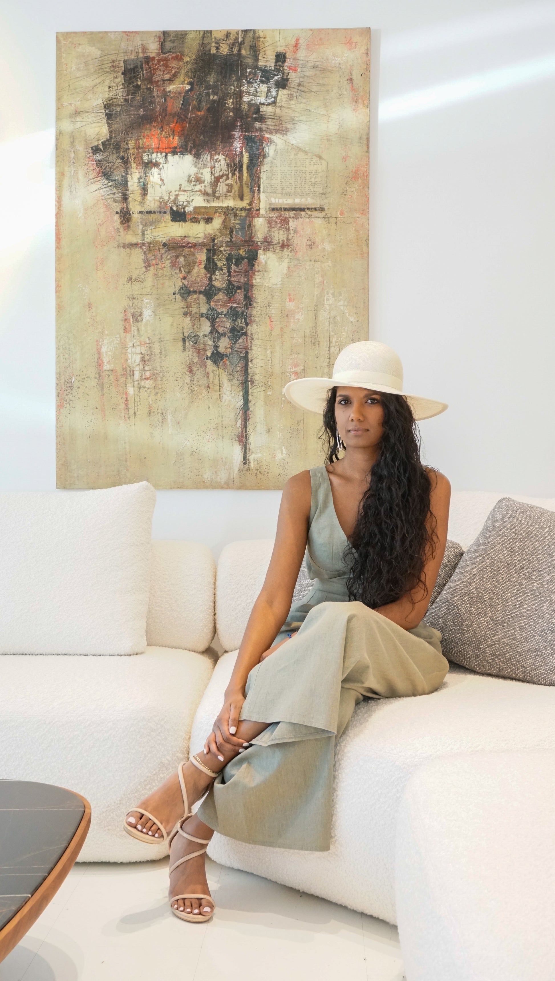 The Hera luxury handcrafted wide-brim ramie-sisal straw hat in champagne, featuring a light cream Italian cotton grosgrain band & a tailored grosgrain bound edge. Shown in a full-length front view on a woman sitting on a couch and looking at the camera. An abstract painting and luxury interior in the background. An exquisite Royal Ascot, race day & couture wedding hat or for everyday luxury. Bespoke Couture Millinery by Anna Xaniotaki