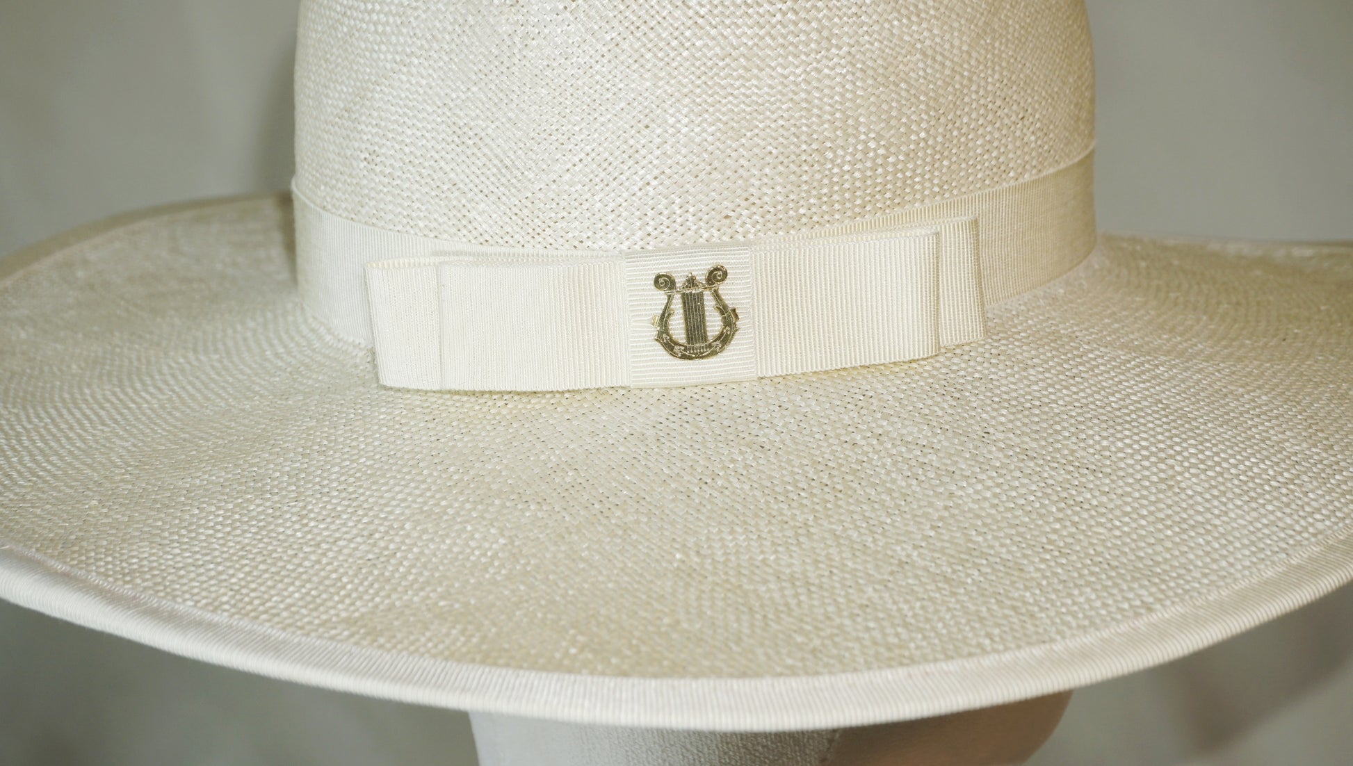 Macro detail shot of The Hera luxury handcrafted wide-brim ramie-sisal straw hat, highlighting the 14ct gold-plated lyre jewel carved with Minoan sea lilies, set on a light cream Italian cotton grosgrain band and bow with a hand-basted and machine-stitched grosgrain bound edge. Bespoke Couture Millinery by Anna Xaniotaki 