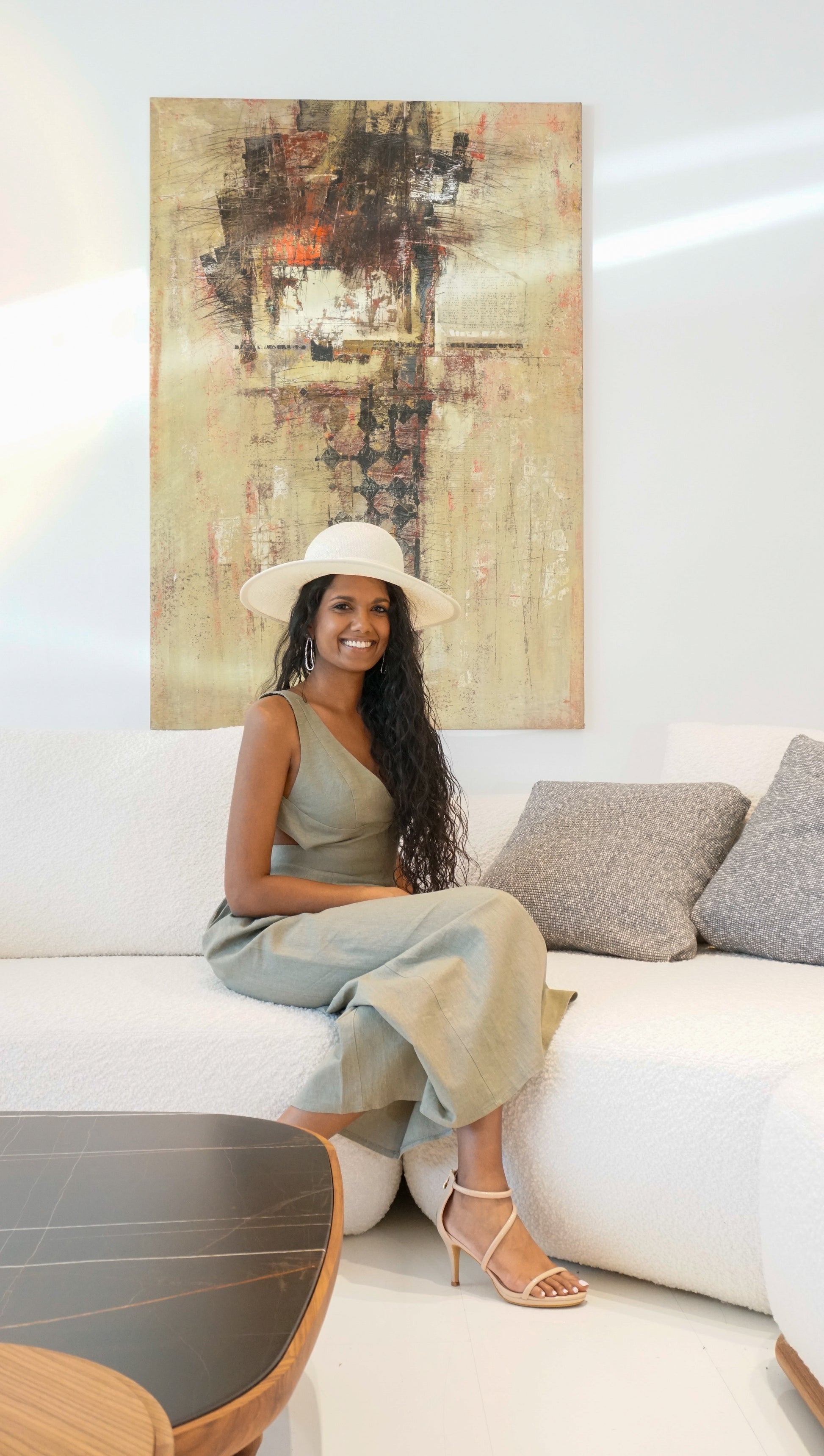 The Hera luxury handcrafted wide-brim ramie-sisal straw hat in champagne, featuring a light cream Italian cotton grosgrain band & a tailored grosgrain bound edge. Shown in a three-quarter length front view on a smiling woman sitting on a white bouclé couch with an abstract painting in the background. An exquisite Royal Ascot, race day & couture wedding hat or for everyday luxury. Bespoke Couture Millinery by Anna Xaniotaki