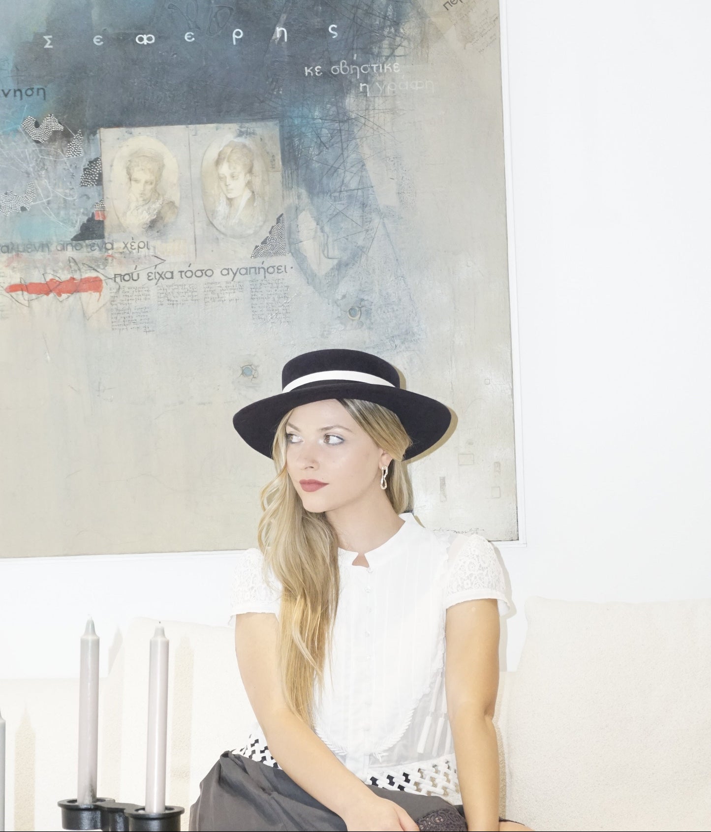 A woman sits on a white couch in a close-up three-quarter front left view, looking away, wearing the handcrafted Kalypso boater hat in luxury navy blue velour rabbit fur felt with an ivory and black Italian cotton grosgrain band. A light blue Seferis artwork frames the background, with two grey-blue candles in the foreground. An exquisite choice for Royal Ascot, race days, cocktail parties and couture weddings, or for everyday luxury elegance. Bespoke Couture Millinery by Anna Xaniotaki