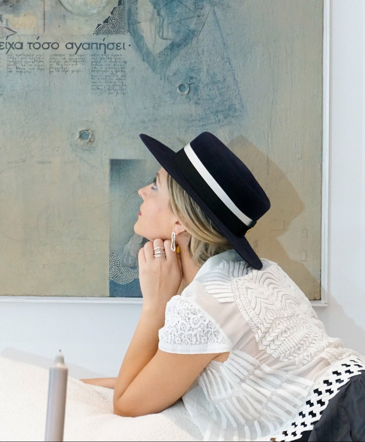 A woman poses elegantly in a left profile view, looking up in front of a light blue artwork while sitting on an ivory couch. She is wearing the handcrafted Kalypso boater hat in luxury navy blue velour rabbit fur felt, which beautifully highlights a black and ivory Italian cotton grosgrain band and the signature 14ct gold-plated sterling silver lyre jewel. An exquisite choice for cocktail parties, art galleries, high-society events or for everyday luxury elegance. Bespoke Couture Millinery by Anna Xaniotaki