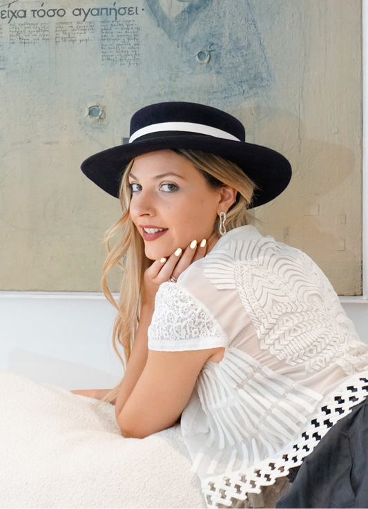 A smiling woman poses elegantly looking over her shoulder while sitting on an ivory couch, wearing the handmade Kalypso boater hat in luxury navy blue velour rabbit fur felt with a black and ivory Italian cotton grosgrain band. Captured in a close-up, left profile view, she rests against the backdrop of a serene light blue artwork. An exquisite choice for race days, weddings, high-society events and for everyday luxury elegance. Bespoke couture millinery by Anna Xaniotaki