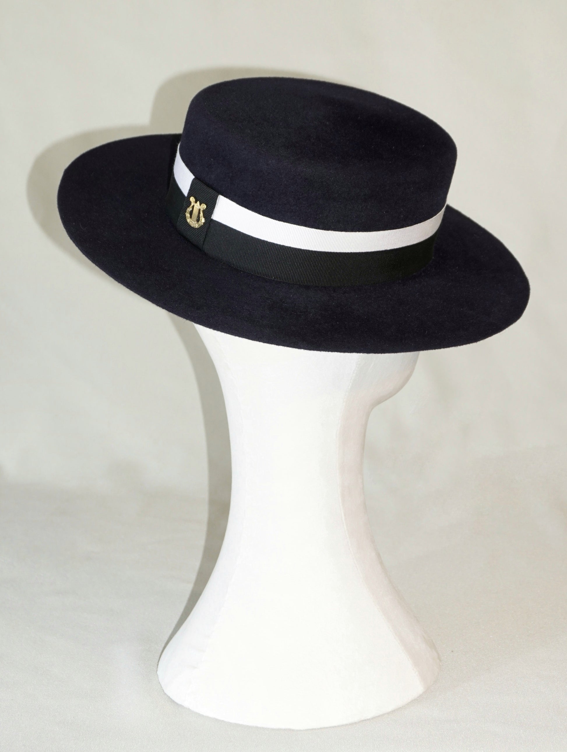 Three-quarter back right view of the handcrafted Kalypso boater hat in luxury navy blue velour rabbit fur felt, displayed on a white mannequin stand showcasing the signature 14ct gold-plated sterling silver lyre jewel and ivory and black Italian cotton grosgrain band. Bespoke Couture Millinery by Anna Xaniotaki
