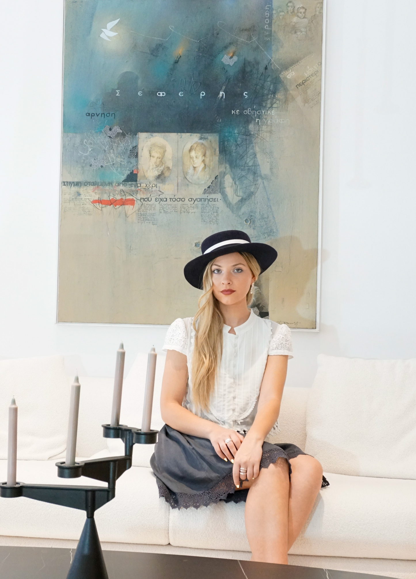 A woman sits elegantly on an ivory couch, wearing the handmade Kalypso boater hat in luxury navy blue velour rabbit fur felt with an ivory and black Italian cotton grosgrain band. Captured in a full-length front view, she showcases the premium couture design. An exquisite choice for race days, weddings, high-society events, or for everyday luxury elegance. Bespoke Couture Millinery by Anna Xaniotaki