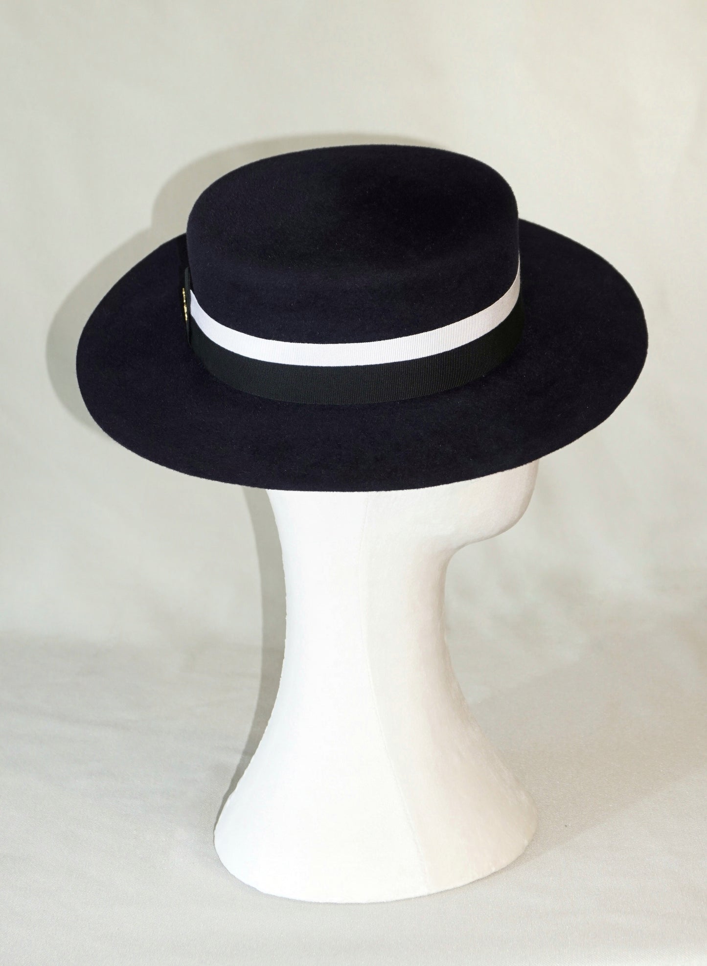 Right profile view of the handcrafted Kalypso boater hat in luxury navy blue velour rabbit fur felt, worn slightly tilted to the right on a white mannequin stand, featuring an ivory and black Italian cotton grosgrain band and the signature 14ct gold-plated sterling silver lyre jewel. Bespoke Couture Millinery by Anna Xaniotaki