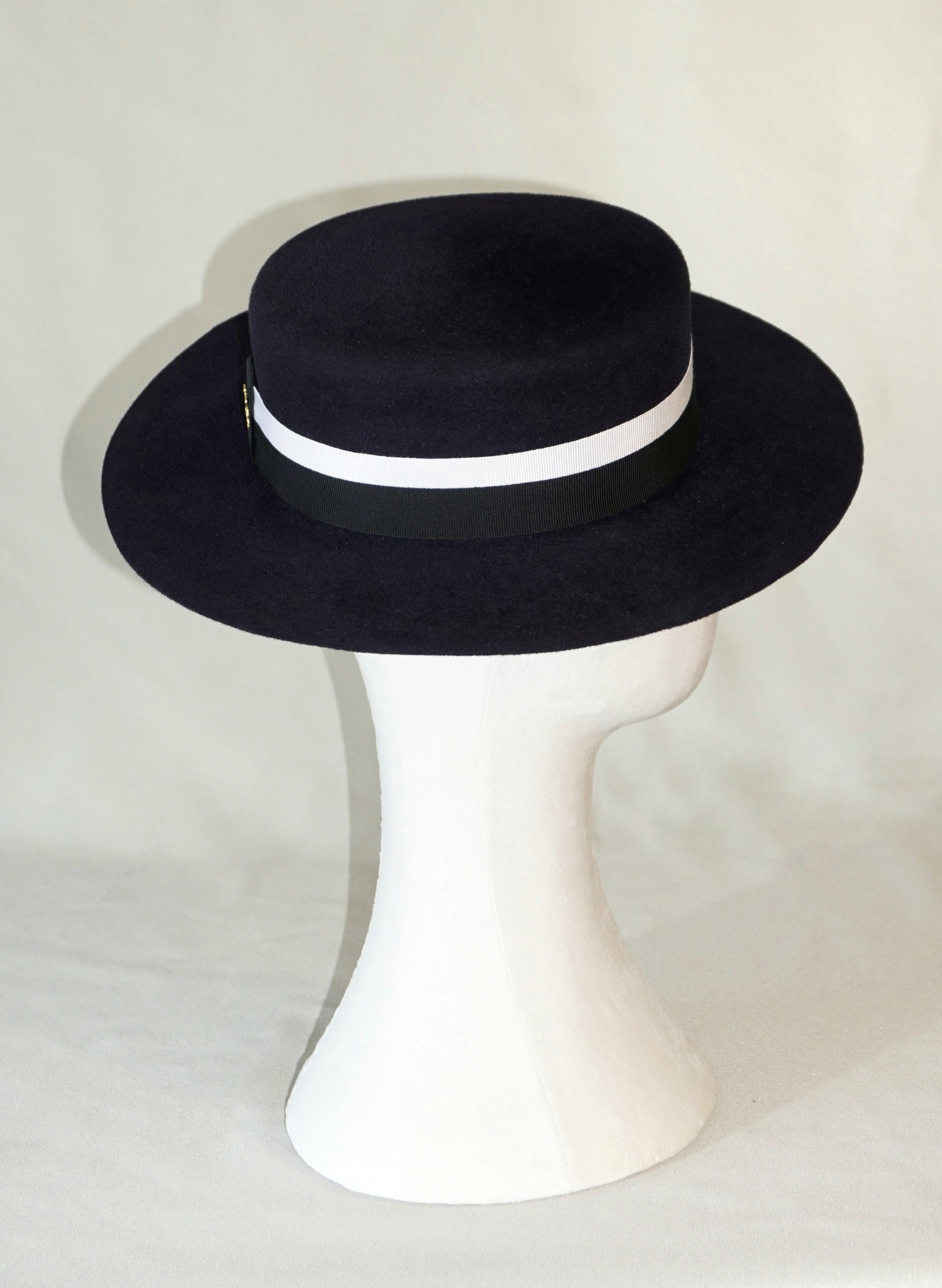 Right profile view of the handcrafted Kalypso boater hat in luxury navy blue velour rabbit fur felt, worn slightly tilted to the right on a white mannequin stand, featuring an ivory and black Italian cotton grosgrain band and the signature 14ct gold-plated sterling silver lyre jewel. Bespoke Couture Millinery by Anna Xaniotaki