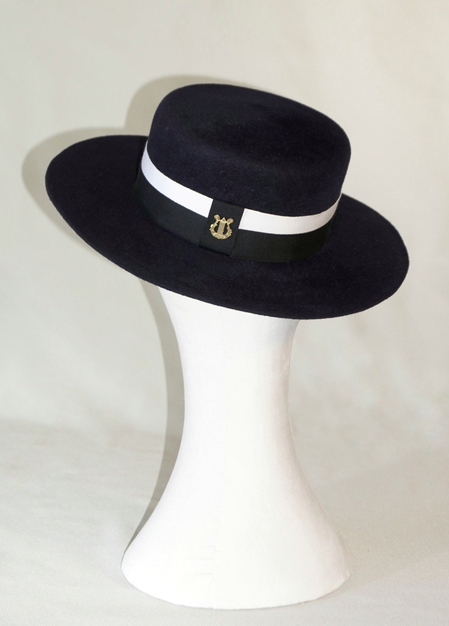 Back view of the handcrafted Kalypso boater hat in luxury navy blue velour rabbit fur felt, worn slightly tilted to the right on a white mannequin stand, showcasing the signature 14ct gold-plated sterling silver lyre jewel and ivory and black Italian cotton grosgrain band. Bespoke Couture Millinery by Anna Xaniotaki