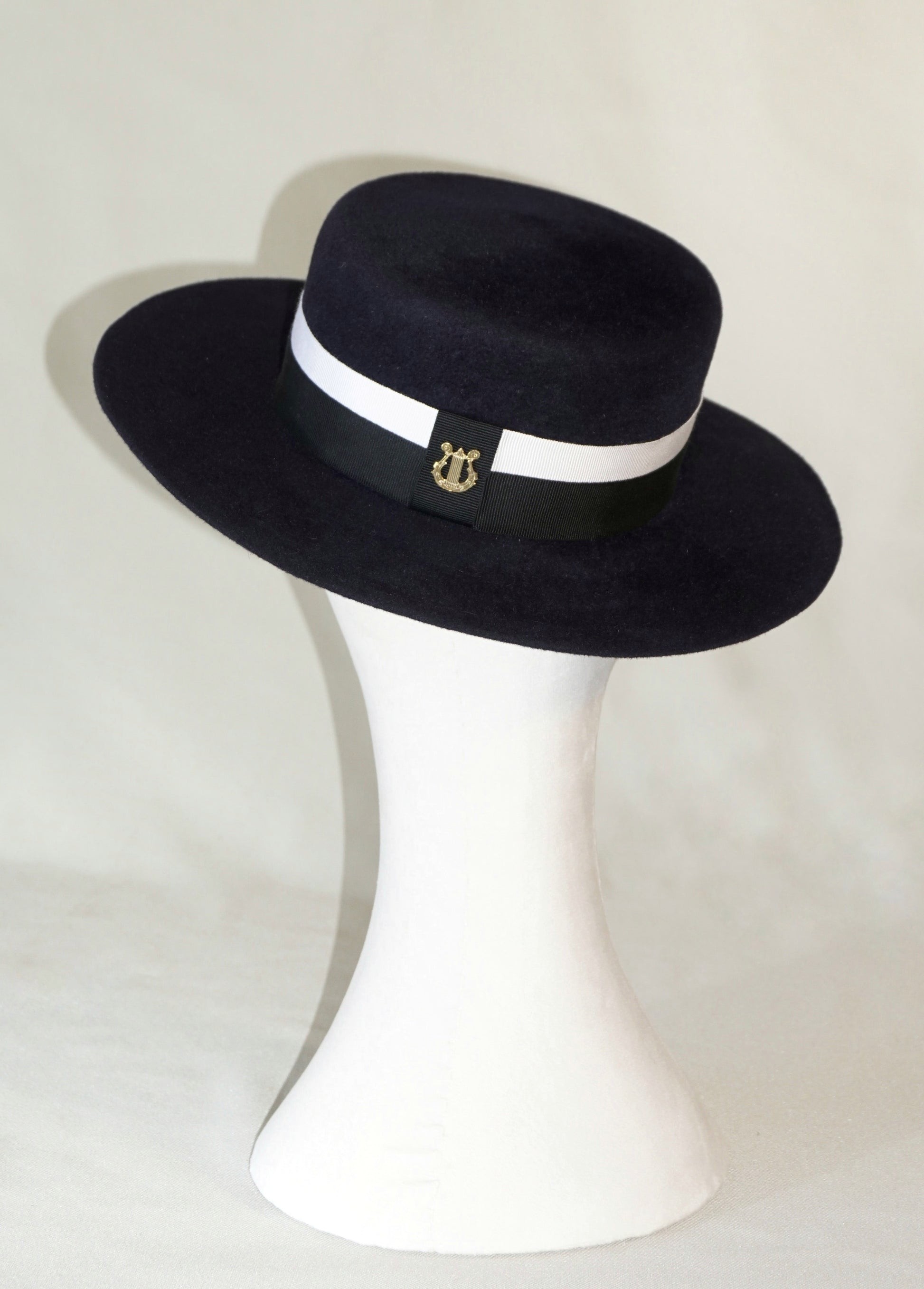 Back view of the handcrafted Kalypso boater hat in luxury navy blue velour rabbit fur felt, worn slightly tilted to the right on a white mannequin stand, showcasing the signature 14ct gold-plated sterling silver lyre jewel and ivory and black Italian cotton grosgrain band. Bespoke Couture Millinery by Anna Xaniotaki
