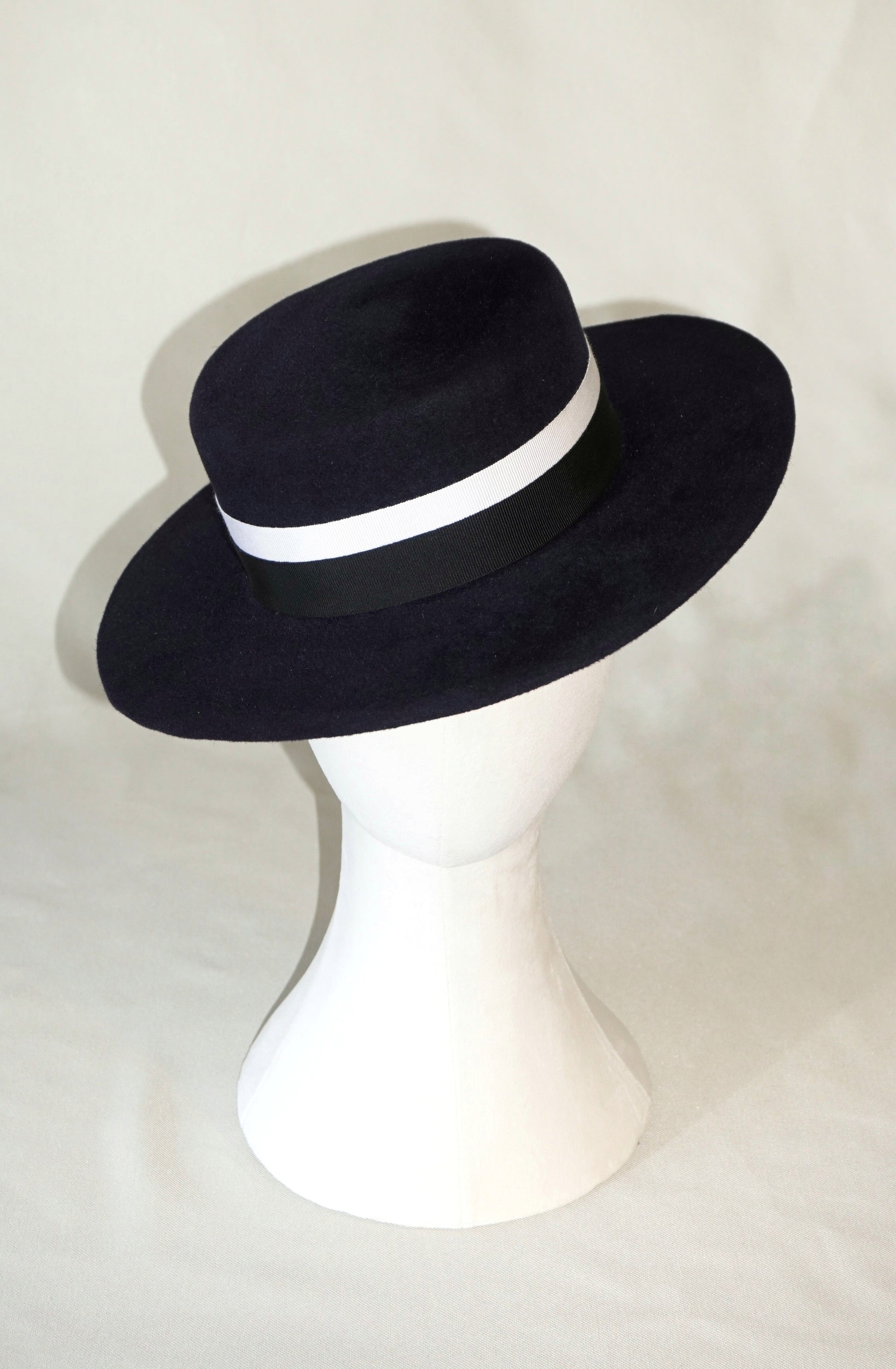Front view of the handcrafted Kalypso boater hat in luxury navy blue velour rabbit fur felt, worn slightly tilted on a white mannequin stand, featuring an ivory and black Italian cotton grosgrain band. Bespoke Couture Millinery by Anna Xaniotaki