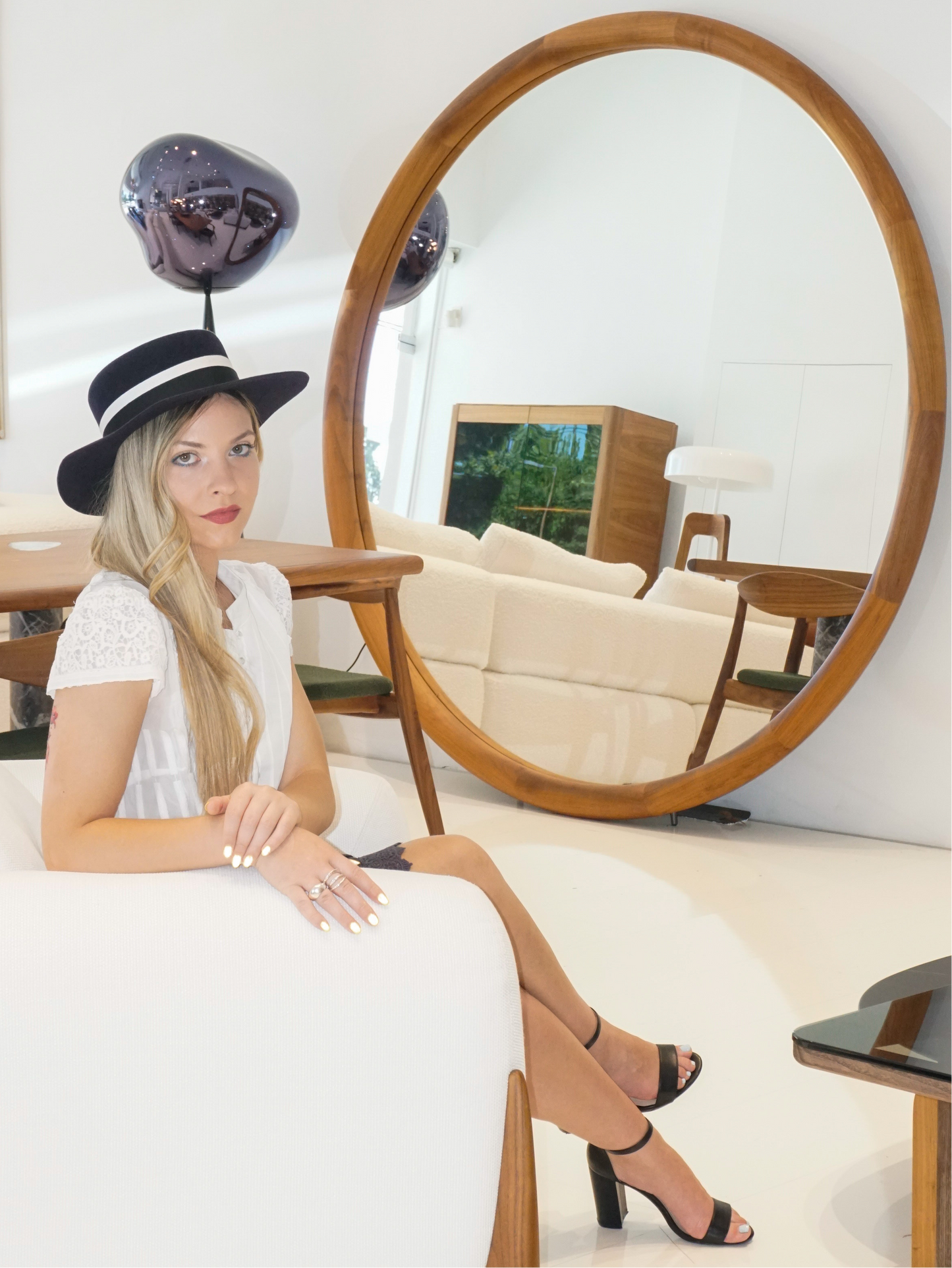 A woman poses elegantly on a white armchair, wearing the handmade Kalypso boater hat in luxury navy blue velour rabbit fur felt with an ivory and black Italian cotton grosgrain band. Captured in a full-length, three-quarter front right view, she showcases the premium couture design. An exquisite choice for race days, weddings, high-society events and for everyday luxury elegance. Bespoke couture millinery by Anna Xaniotaki