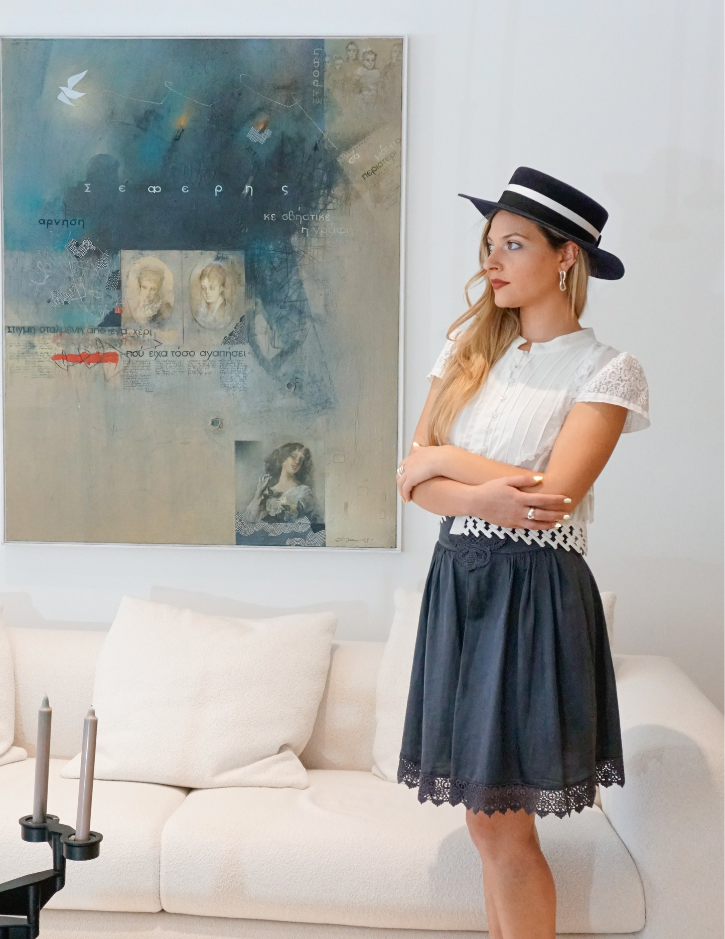 A woman stands in a three-quarter length left profile view wearing the handcrafted Kalypso boater hat in luxury navy blue velour rabbit fur felt with an ivory and black grosgrain band. A light blue Seferis artwork and white couch frame the background. An exquisite choice for Royal Ascot, race days, cocktail parties and couture weddings, or for everyday luxury elegance. Bespoke Couture Millinery by Anna Xaniotaki