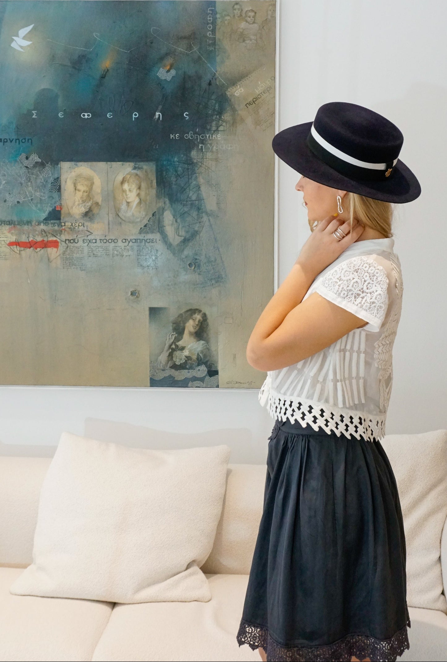 A woman stands in a left side view, showcasing the handcrafted Kalypso boater hat in luxury navy blue velour rabbit fur felt with an ivory and black grosgrain band and a 14ct gold-plated sterling silver lyre jewel. A light blue Seferis artwork and white couch frame the background. An exquisite choice for cocktail parties, art galleries, high-society events or for everyday luxury elegance. Bespoke Couture Millinery by Anna Xaniotaki
