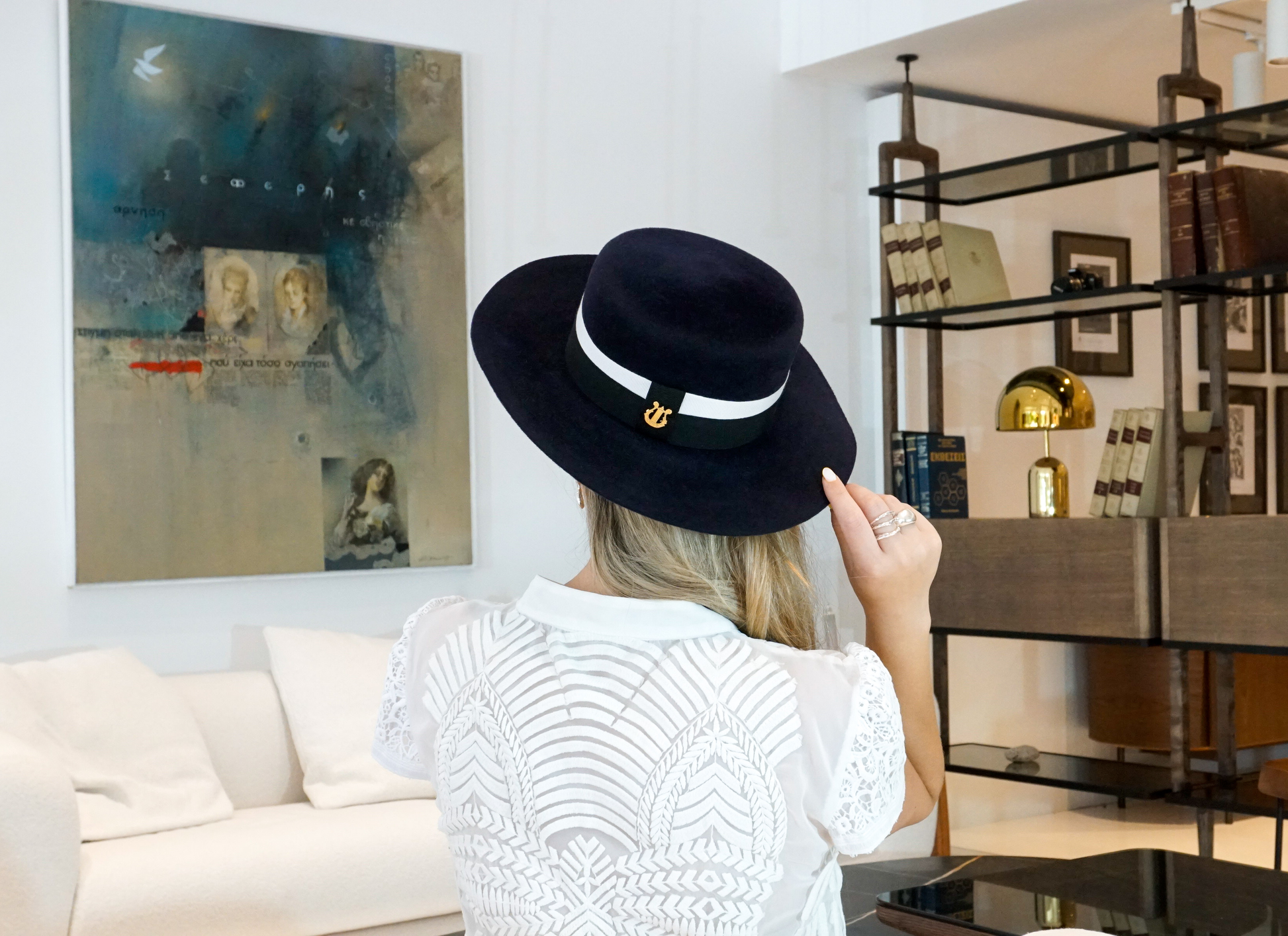 A close-up back view of a woman showcasing the signature 14ct gold-plated sterling silver lyre jewel on the handcrafted Kalypso boater hat in luxury navy blue velour rabbit fur felt with an ivory and black grosgrain band. A light blue Seferis artwork, white couch, and modern wooden bookshelf frame the background. An exquisite choice for cocktail parties, art galleries, high-society events or for everyday luxury elegance. Bespoke Couture Millinery by Anna Xaniotaki