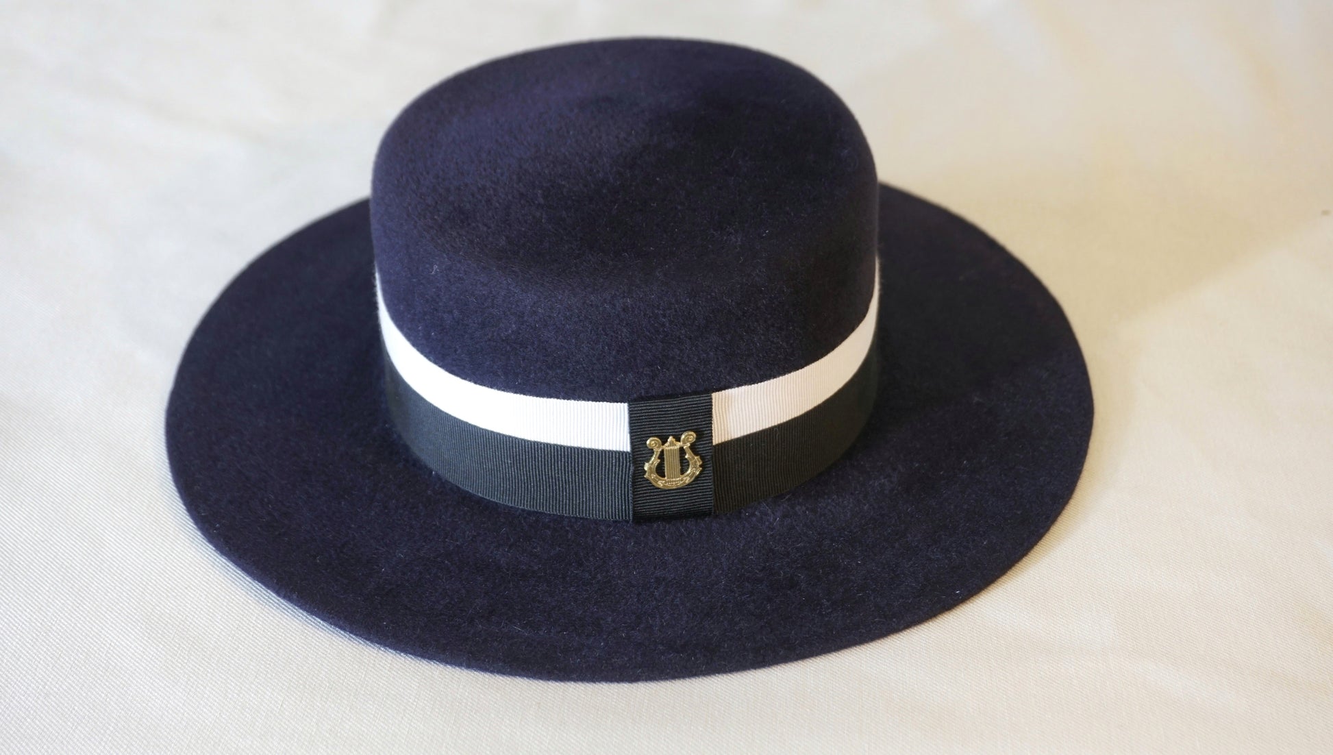 Product shot of the handcrafted Kalypso boater hat in luxury navy blue velour rabbit fur felt, back view showcasing the signature 14ct gold-plated sterling silver lyre jewel and ivory and black Italian cotton grosgrain band against a neutral background. Bespoke Couture Millinery by Anna Xaniotaki