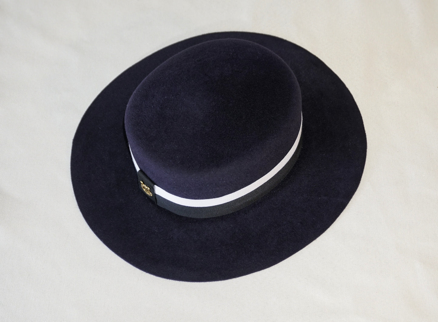 High-angle top view product shot of the handcrafted Kalypso boater hat in luxury navy blue velour rabbit fur felt, revealing the flat crown, ivory and black Italian cotton grosgrain band, and the signature 14ct gold-plated sterling silver lyre jewel against a neutral background. Bespoke Couture Millinery by Anna Xaniotaki.