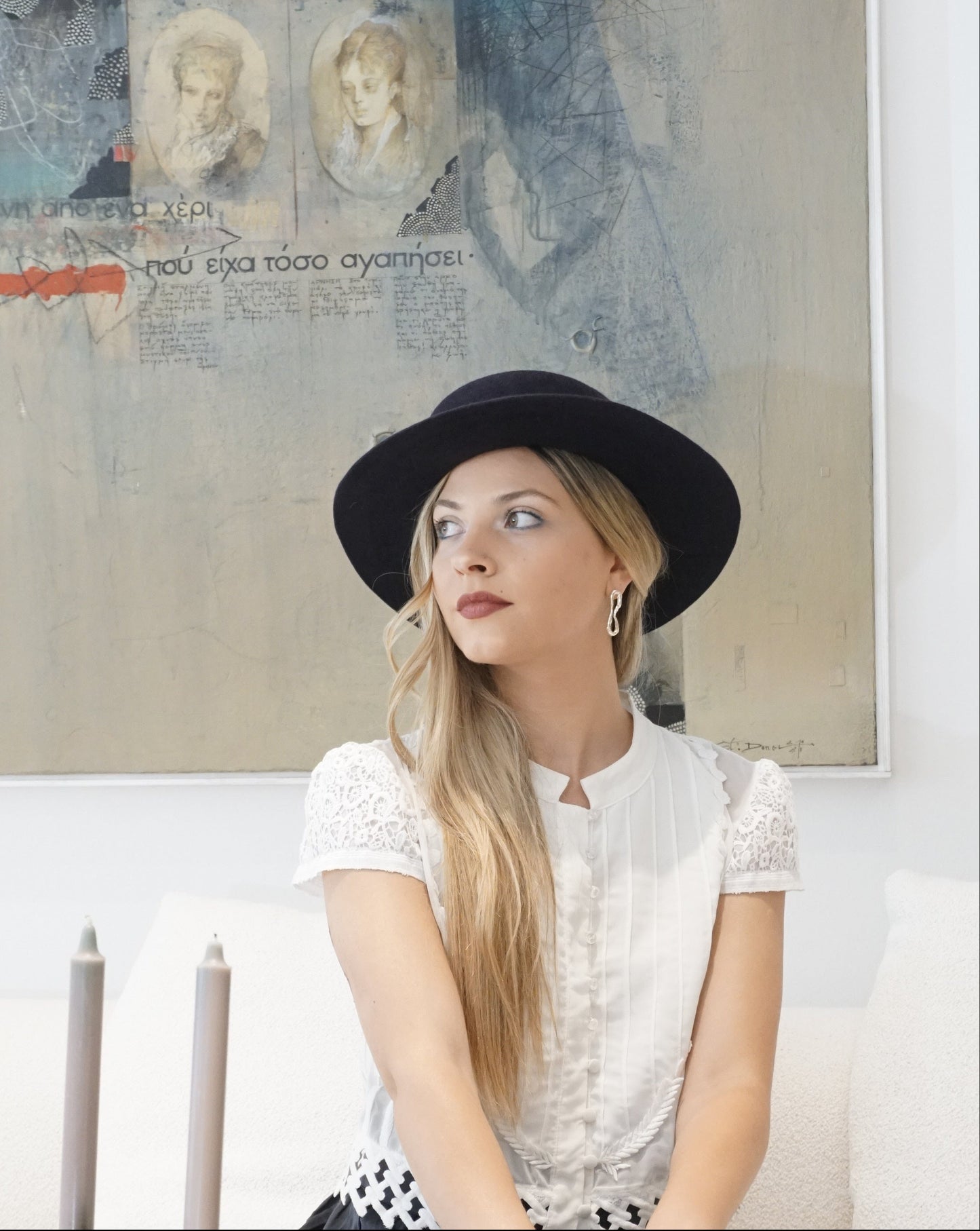A woman poses elegantly in a three-quarter front view, looking right in front of a light blue artwork while sitting on an ivory couch. She is wearing the handcrafted Kalypso boater hat in luxury navy blue velour rabbit fur felt. An exquisite choice for Royal Ascot, race days, cocktail parties and couture weddings, or for everyday luxury elegance. Bespoke Couture Millinery by Anna Xaniotaki
